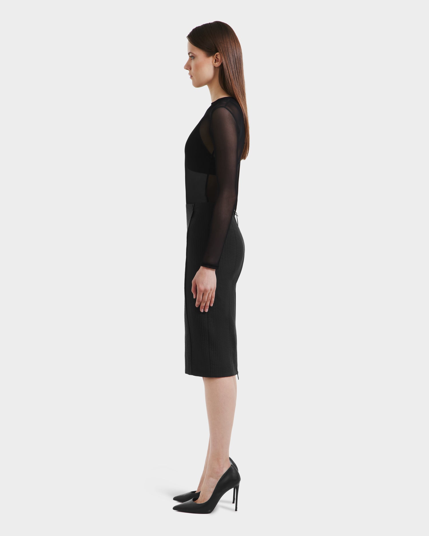A woman stands in profile against a plain background, wearing the Rudsak GLENDORA - 8525563 Black fitted skirt with sheer long sleeves and black high heels. Her straight brown hair is worn down.