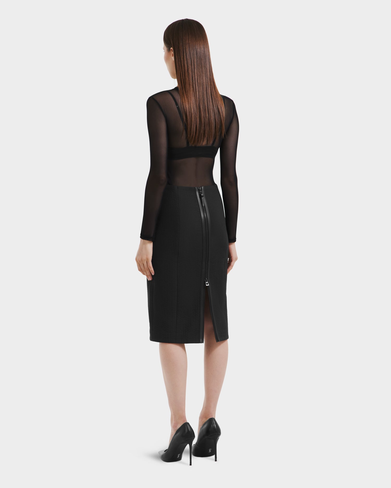 A woman with straight brown hair stands facing away, wearing a sheer black top, black high heels, and the Rudsak GLENDORA - 8525563 Black womens fitted vegan leather midi skirt with a visible back zipper.