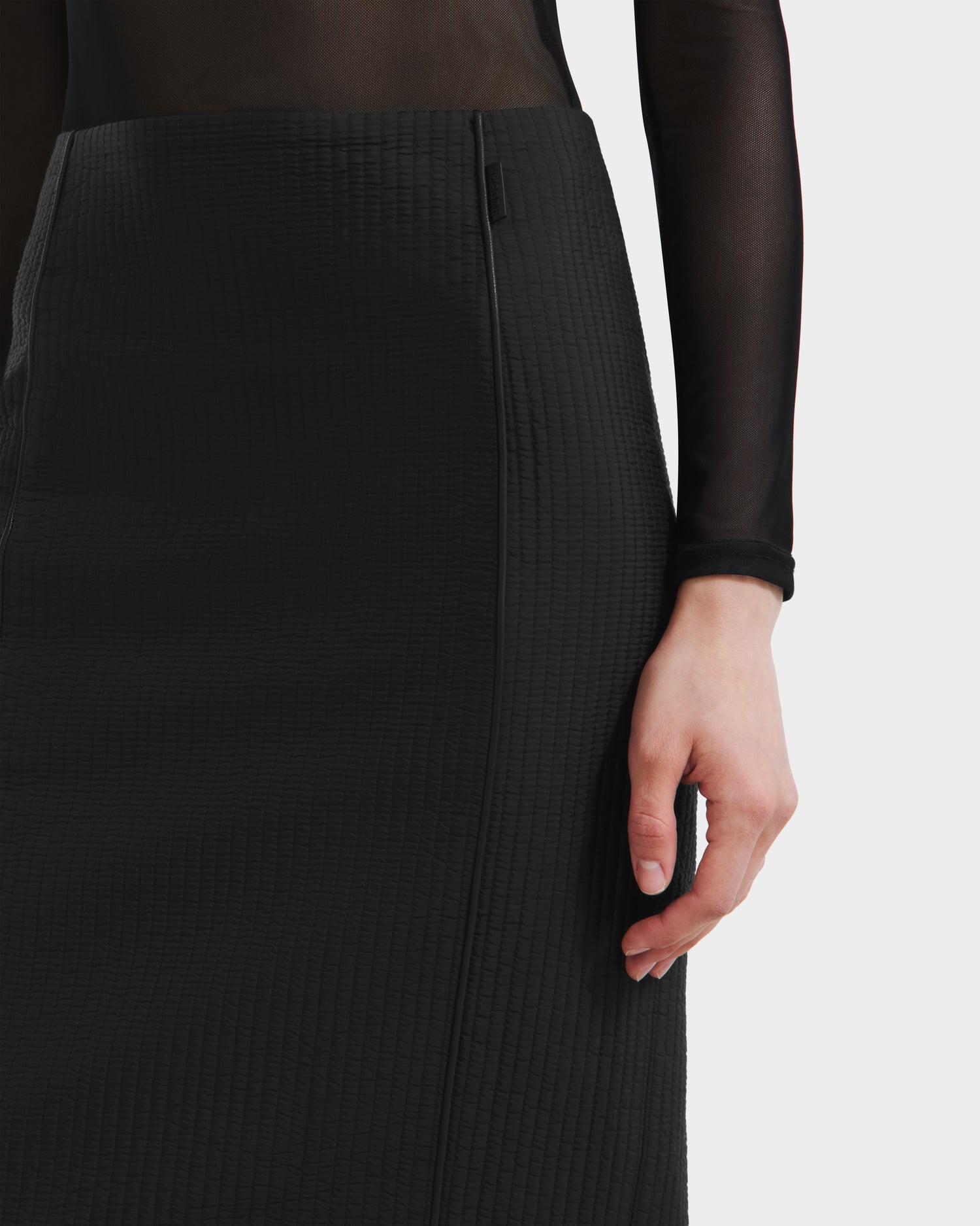 Wearing the Rudsak GLENDORA - 8525563 Black: a sheer long-sleeve top paired with a textured high-waisted vegan leather midi skirt, one arm relaxed at their side against a plain light background.