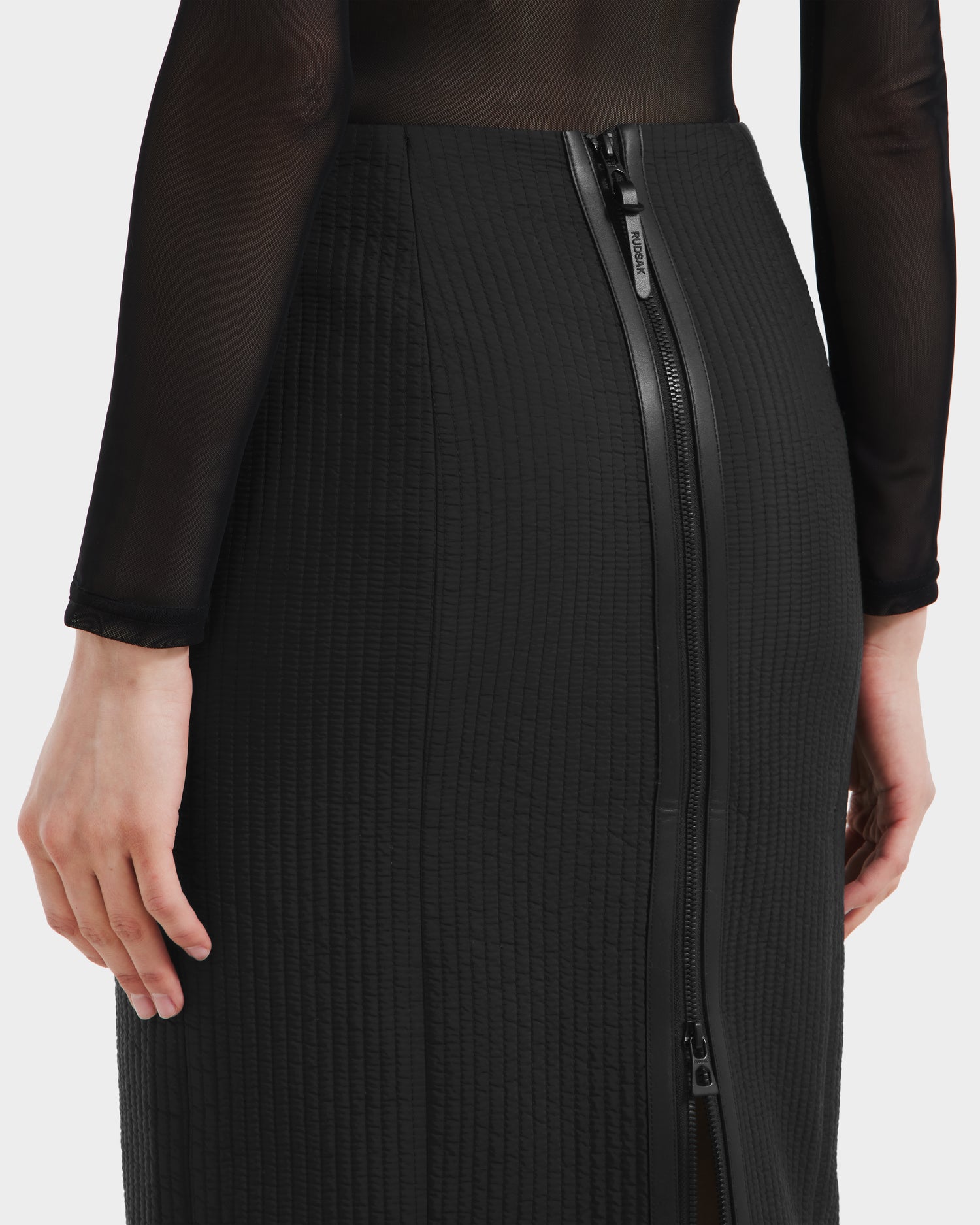 Wearing the Rudsak GLENDORA - 8525563 Black textured vegan leather midi skirt with an exposed back zipper, paired with a sheer long-sleeve top, she creates a sleek, modern look as her hand rests by her side.