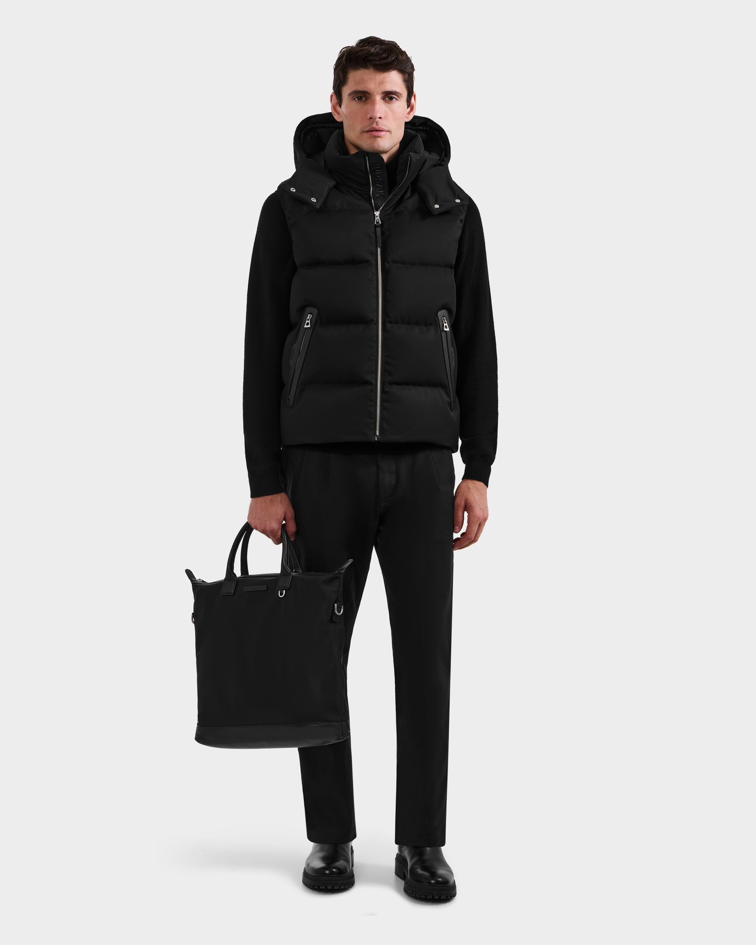 A man stands against a plain background wearing the Rudsak HARLOW - M125528 Black men’s quilted down vest with removable hood, black shirt, pants, and shoes, holding a black tote bag in his left hand.
