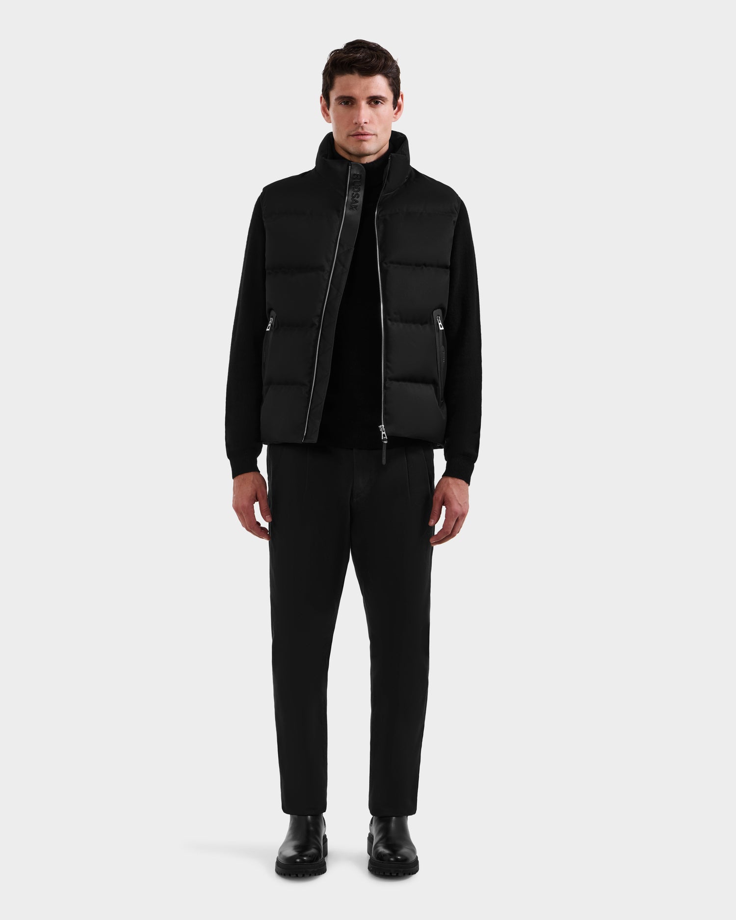 A man faces forward against a plain background, wearing Rudsak’s HARLOW - M125528 Black men’s quilted down vest with removable hood, paired with a black long-sleeve shirt, black trousers, and black boots. His arms are relaxed at his sides.