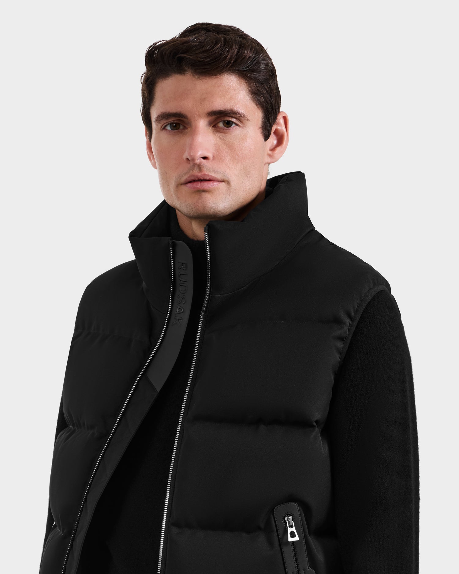 A man with short brown hair wears the Rudsak HARLOW - M125528 Black men’s quilted down puffer vest with a removable hood over a black top, facing forward with a neutral expression against a plain light gray background.