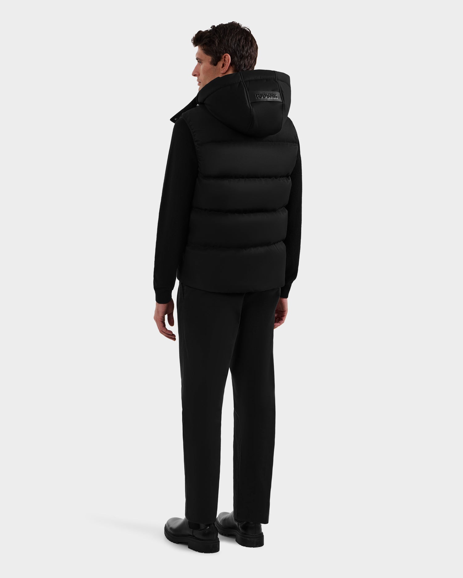 A man stands facing away, wearing the Rudsak HARLOW - M125528 Black men's quilted down vest with removable hood, paired with black pants and shoes against a plain white background.