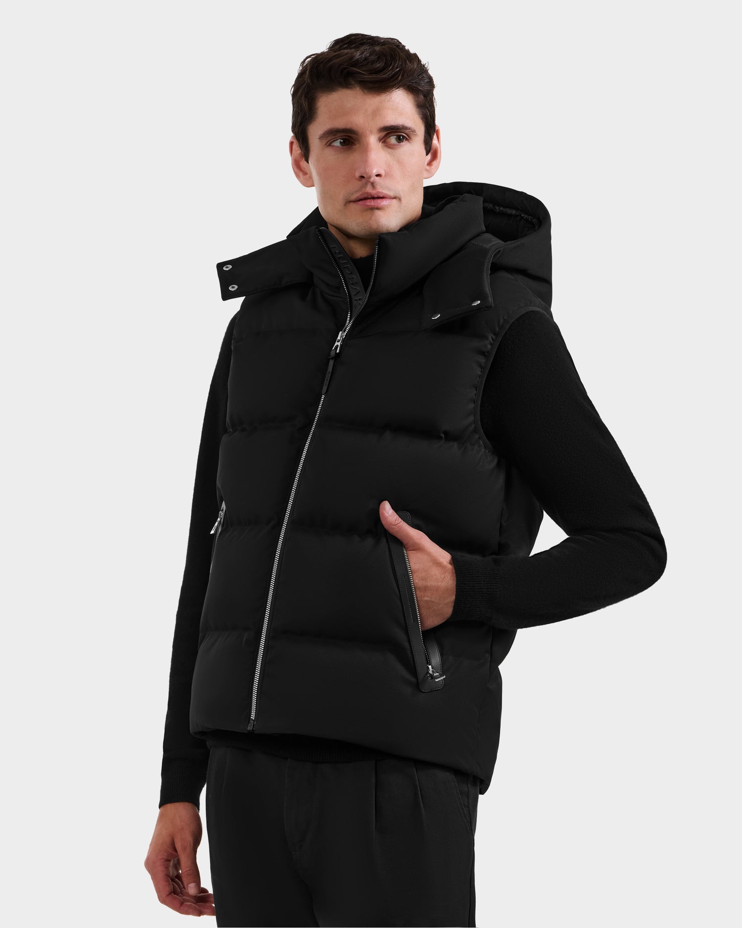 A man wearing the Rudsak HARLOW - M125528 Black quilted down vest with removable hood over a black long-sleeve shirt stands with one hand in his vest pocket, looking slightly to the side against a plain light background.