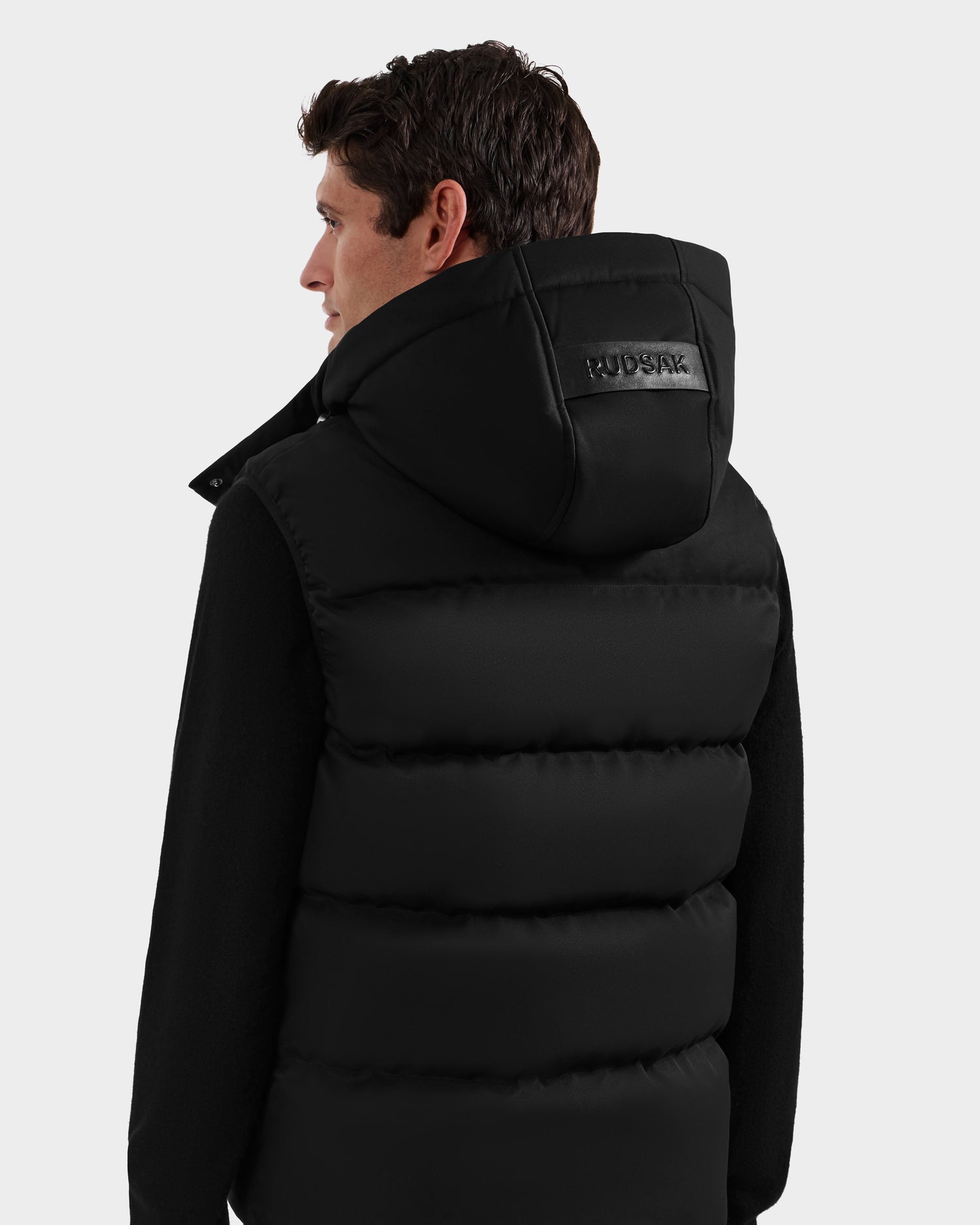 A man with short dark hair, seen from behind, wears the Rudsak HARLOW - M125528 Black men's quilted down vest with a removable hood and a leather RUDSAK patch on the back. The background is plain light gray.