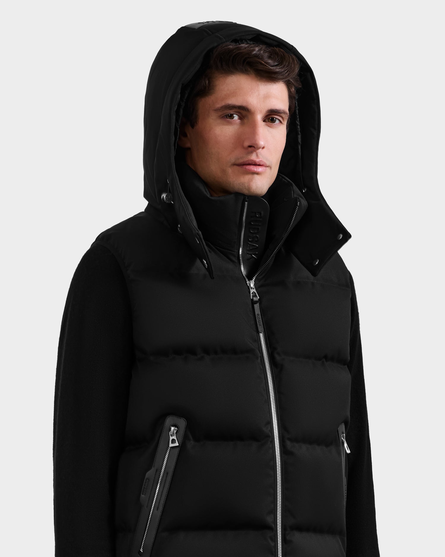 A man wears the Rudsak HARLOW - M125528 Black men’s quilted down vest with a removable hood, silver zippers, and high collar over a black zip-up jacket. He looks at the camera with a neutral expression against a plain light gray background.