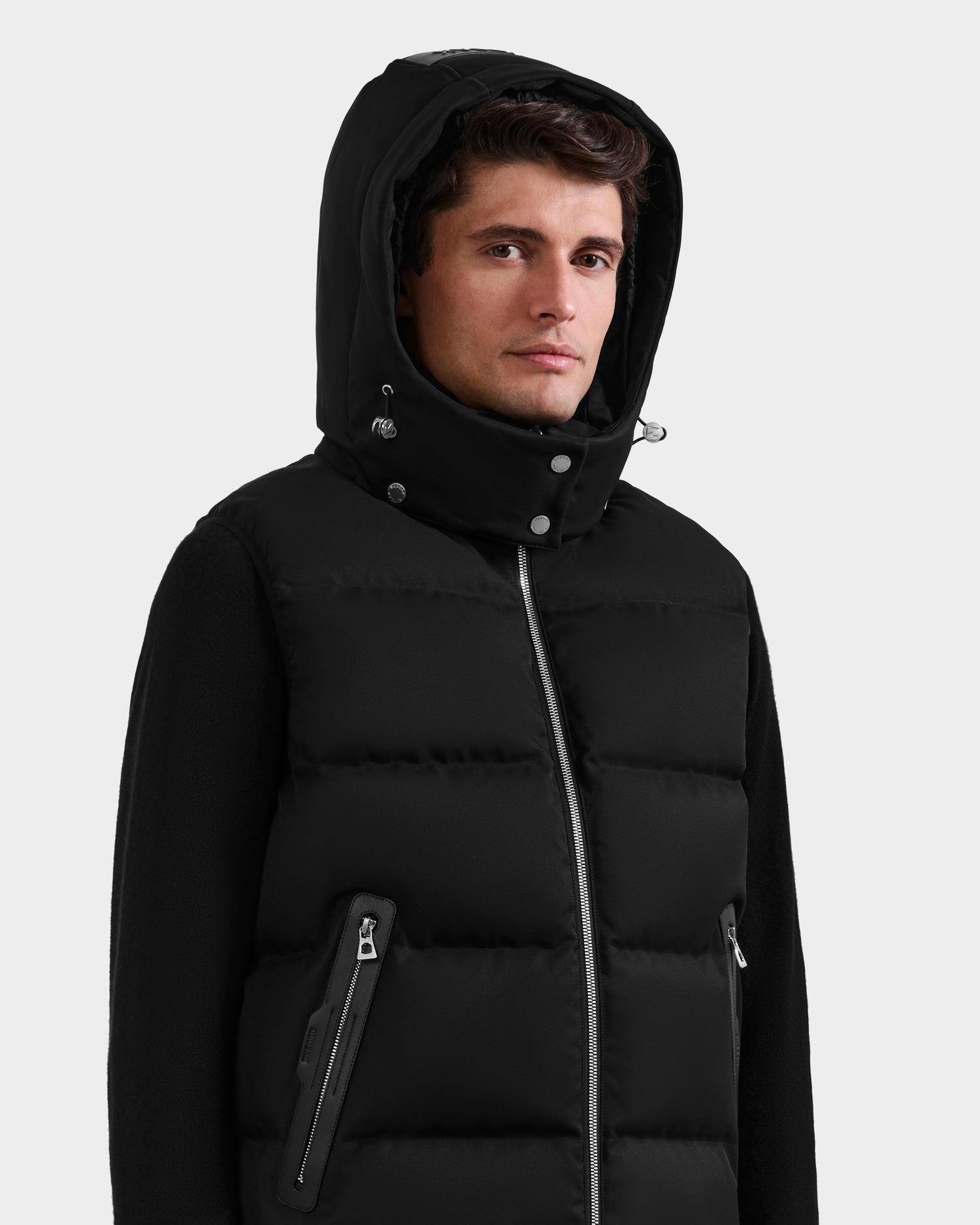 A man stands against a plain light gray background wearing the Rudsak HARLOW - M125528 black men’s quilted down vest featuring a front zipper, zippered pockets, and a removable hood.