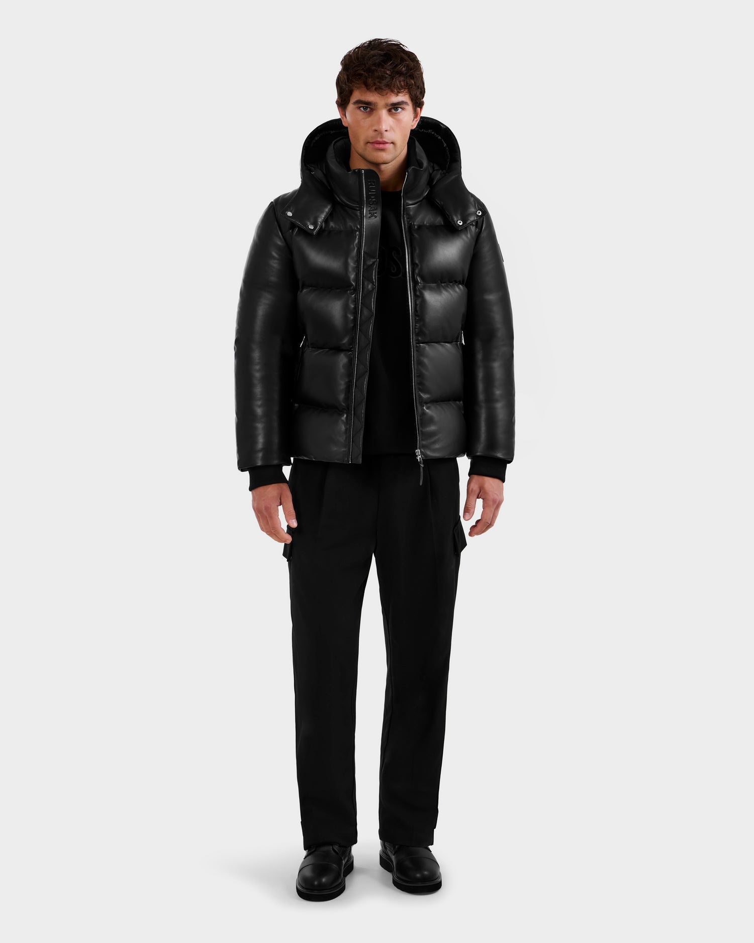A man stands facing forward wearing the Rudsak HARVEY X - 6125681 Black puffer jacket with a hood, black pants, and black shoes against a plain light-gray background.