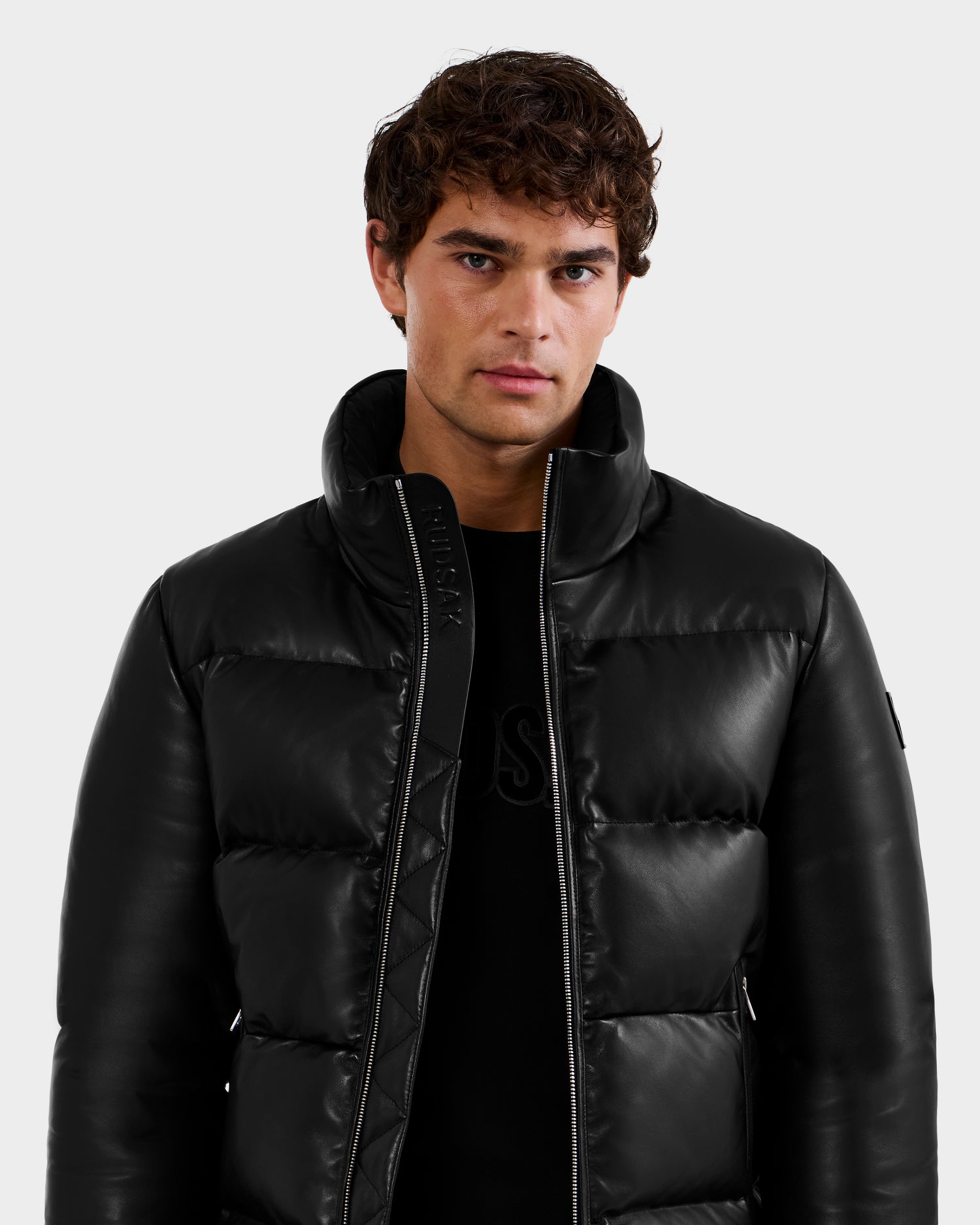 A young man with curly brown hair wears the Rudsak HARVEY X - 6125681 Black puffer jacket over a black shirt, standing against a plain light gray background.