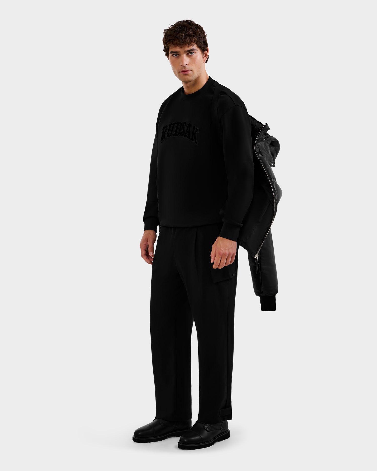 A man stands against a plain background wearing the Rudsak HARVEY X - 6125681 Black sweatshirt with “RUDSAK” on the chest, matching black pants, black shoes, and holding a black jacket over his shoulder.