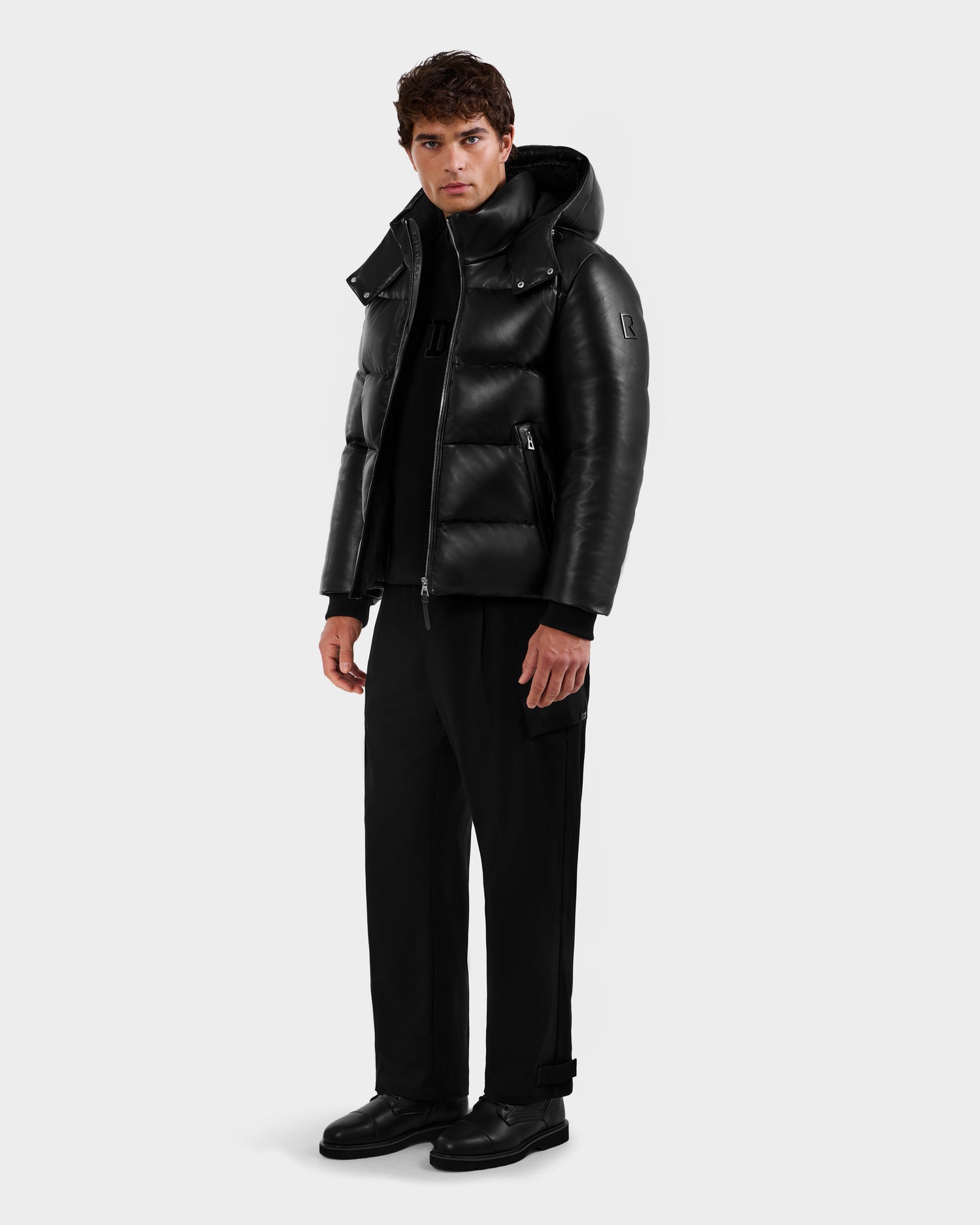 A man faces forward against a plain light background, wearing the Rudsak HARVEY X - 6125681 Black puffer jacket with a high collar and hood, paired with black pants, shoes, and a black turtleneck.