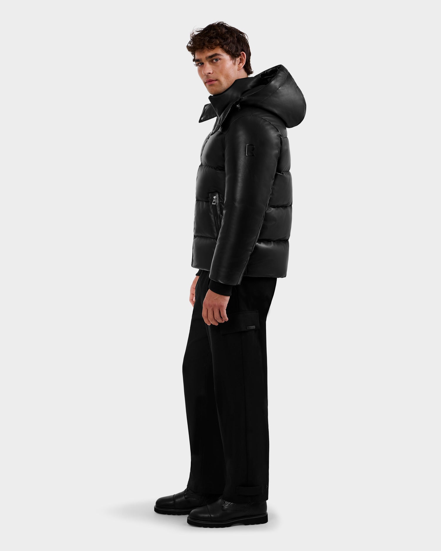A man stands slightly to the side against a plain light background, wearing Rudsak's HARVEY X - 6125681 Black puffer jacket with a hood, paired with black pants and black boots.