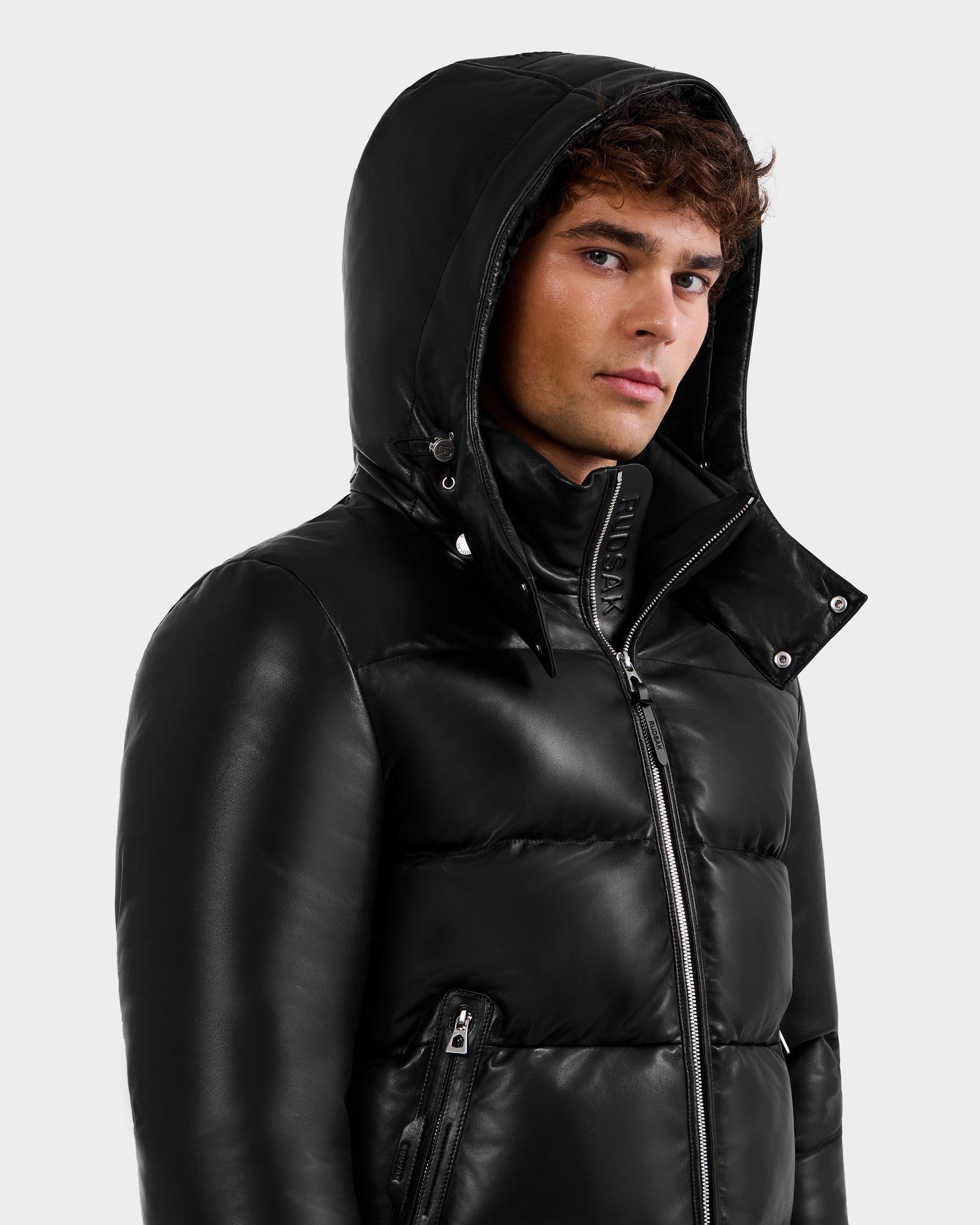 A young man with curly brown hair wears the Rudsak HARVEY X - 6125681 Black puffer jacket with a hood and zippered pockets, looking slightly to the side against a plain light background.