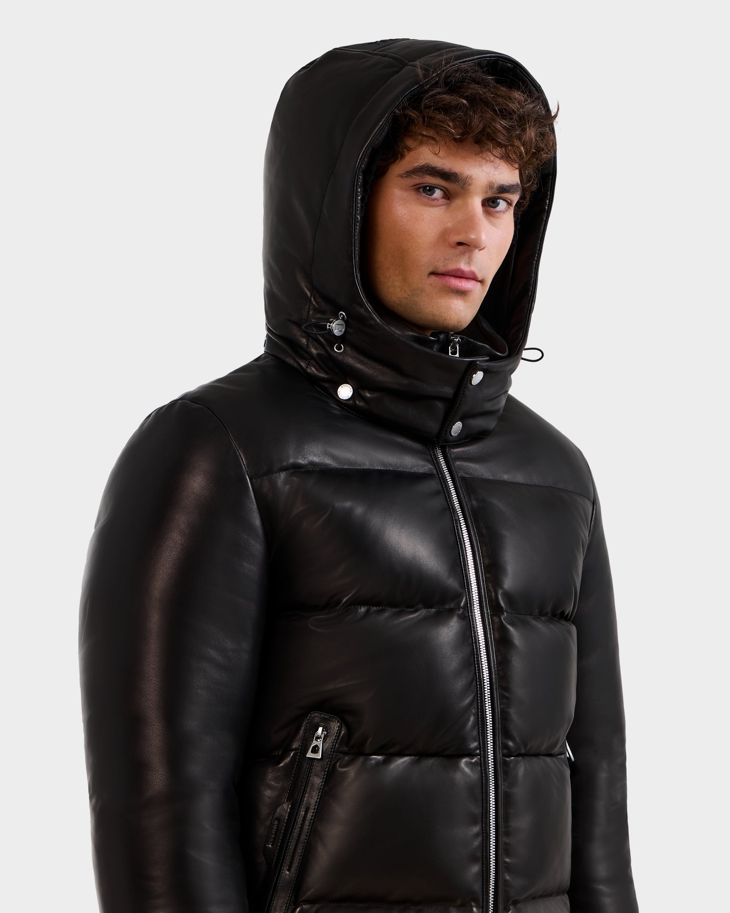 A young man with curly hair wears the Rudsak HARVEY X - 6125681 Black puffer jacket with the hood up, looking slightly to the side against a plain light background.