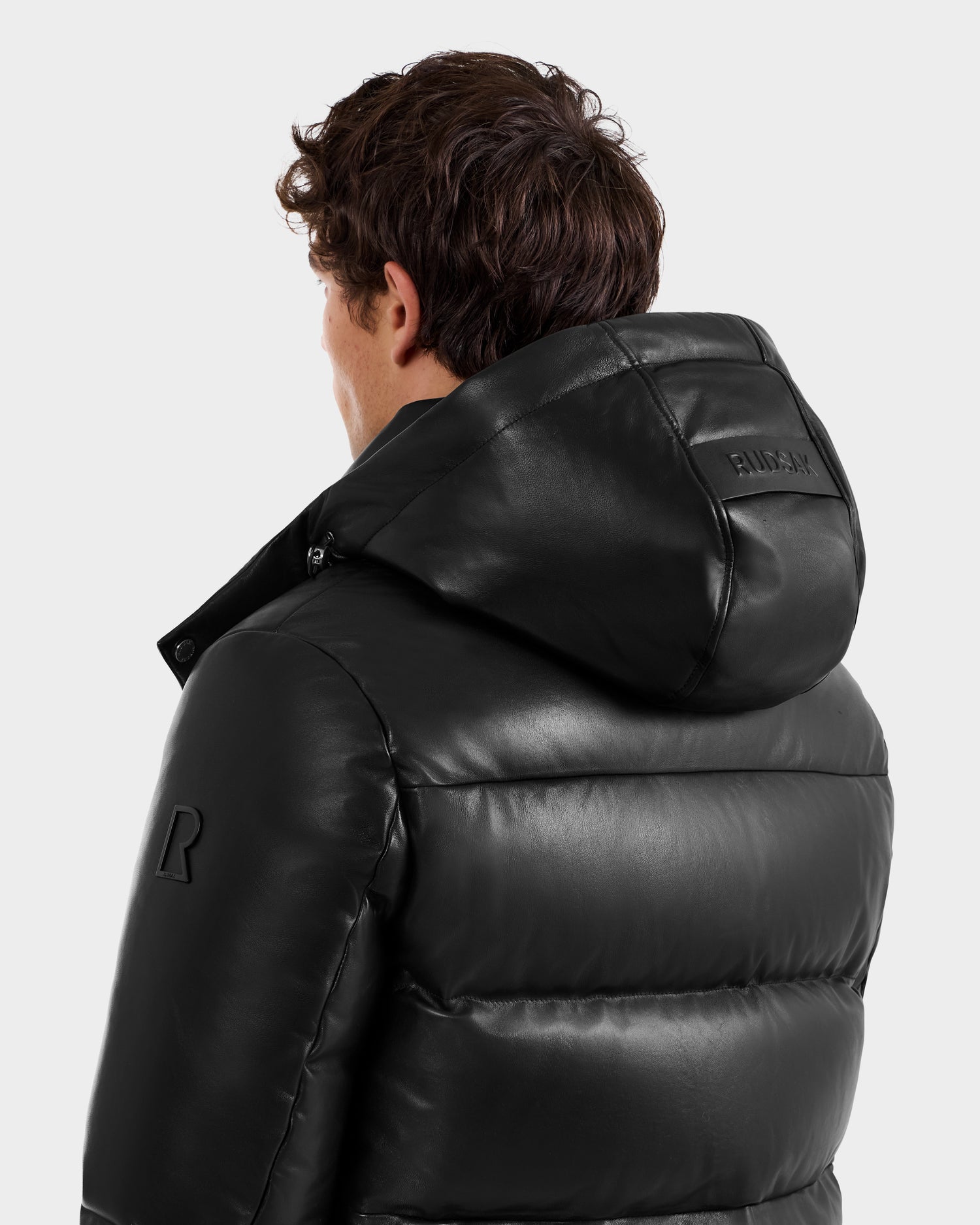 A person with short, curly brown hair is seen from behind wearing the shiny black HARVEY X - 6125681 puffer jacket from Rudsak, with a seam and embossed text on the hood. The background is plain light gray.