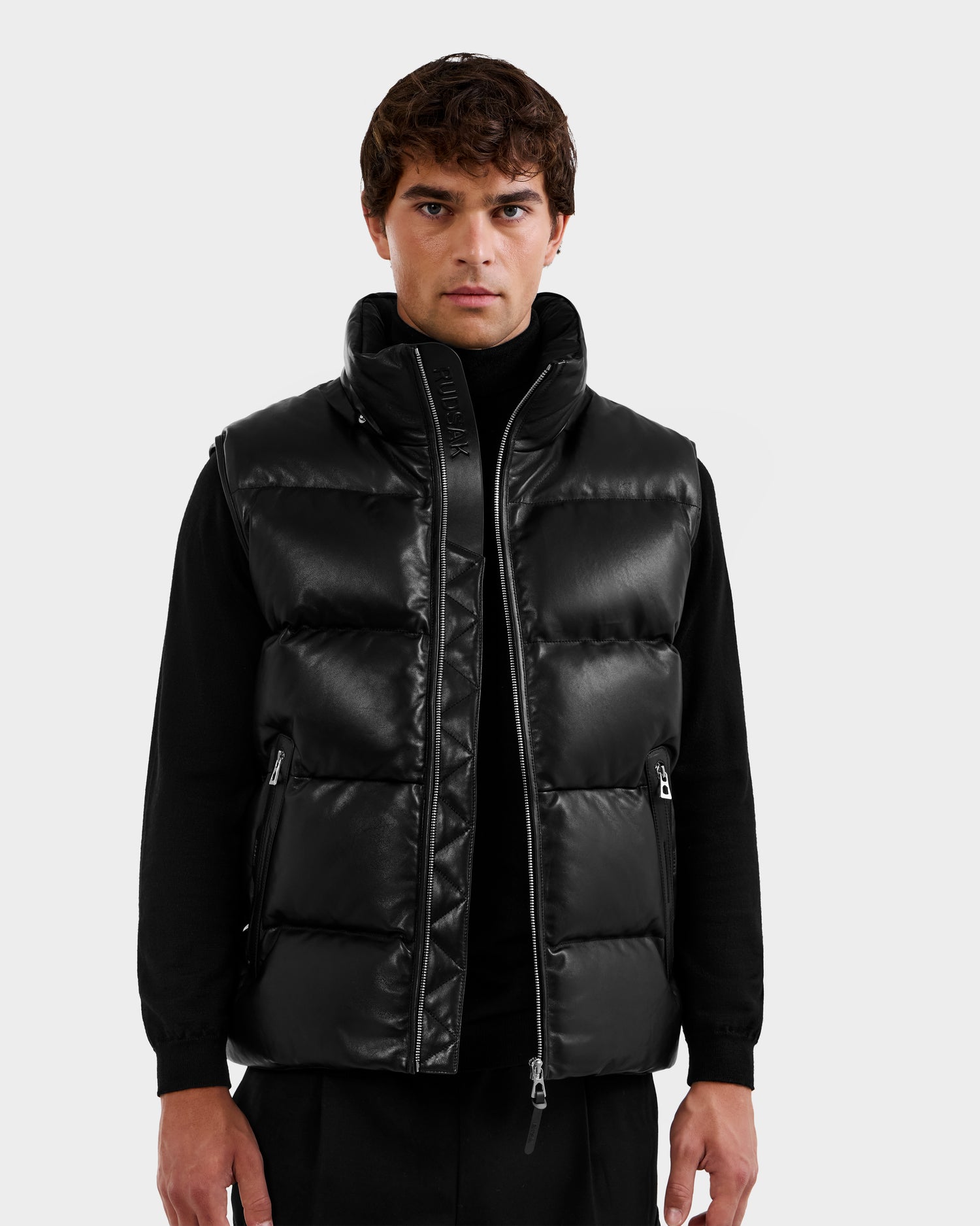 A man wears the Rudsak HENRY X - 6125679 Black puffer vest over a black turtleneck, standing against a plain light gray background and looking directly at the camera.