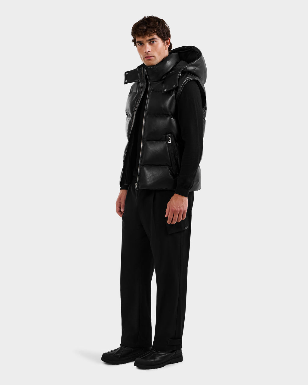 A man faces slightly left, wearing the Rudsak HENRY X - 6125679 Black puffer vest over a black long-sleeve shirt, with black pants and shoes, set against a plain light gray background.