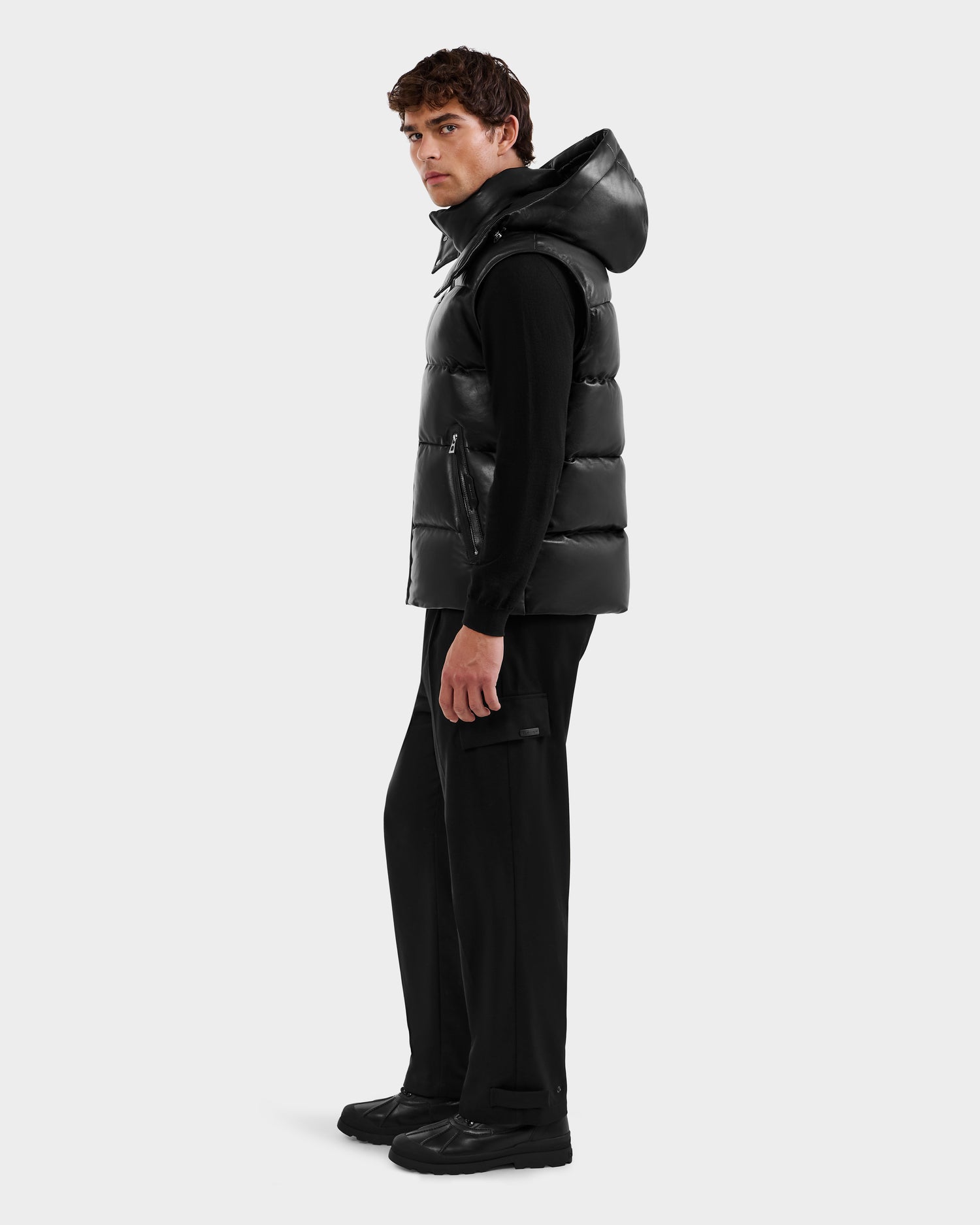 A man stands slightly sideways, wearing the HENRY X - 6125679 Black puffer vest by Rudsak over a black long-sleeve shirt, black pants, and black shoes, set against a plain white background.