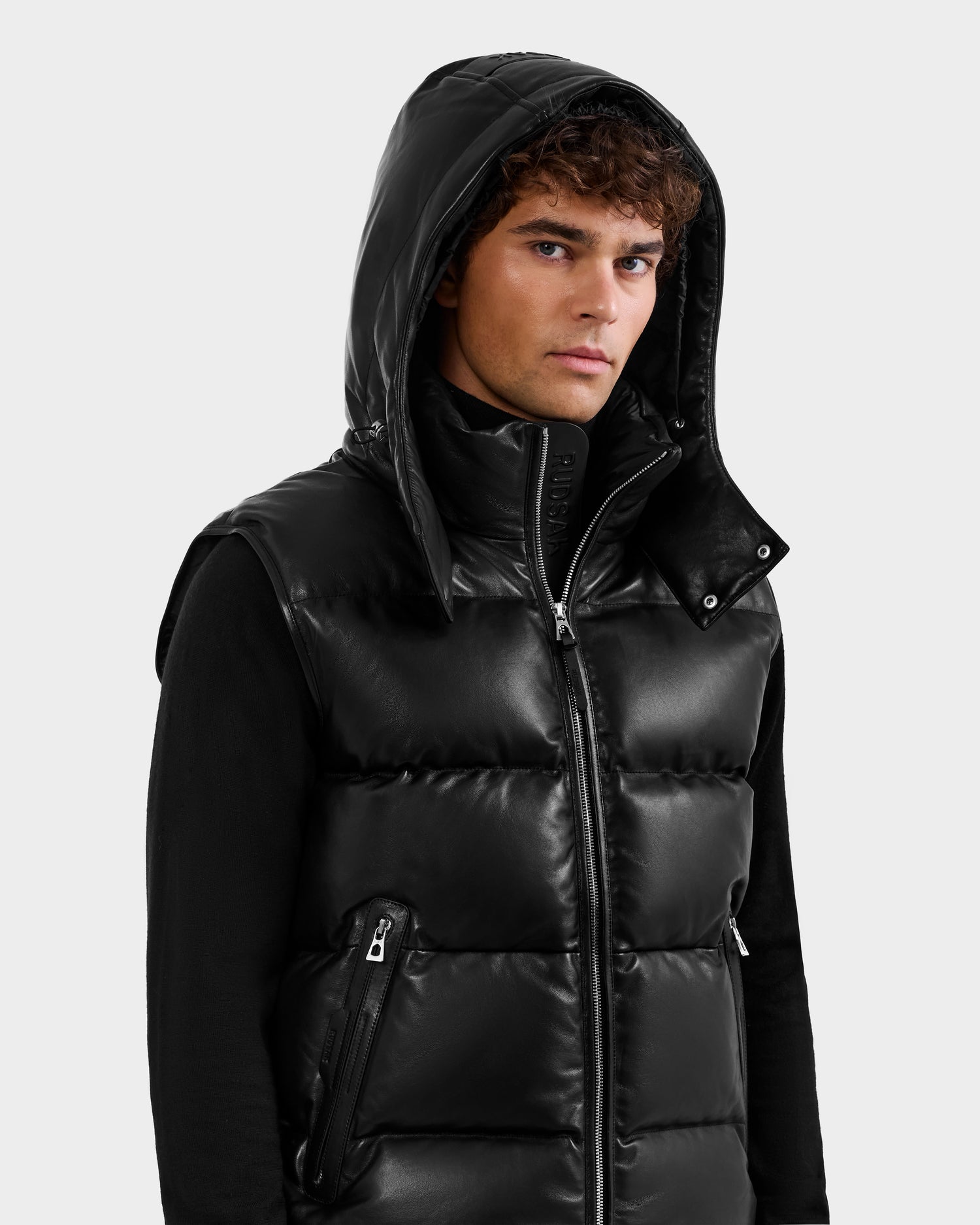 A young man with curly hair wears the Rudsak HENRY X - 6125679 Black shiny hooded puffer vest over a black long-sleeve shirt, facing forward with a neutral expression against a plain light background.