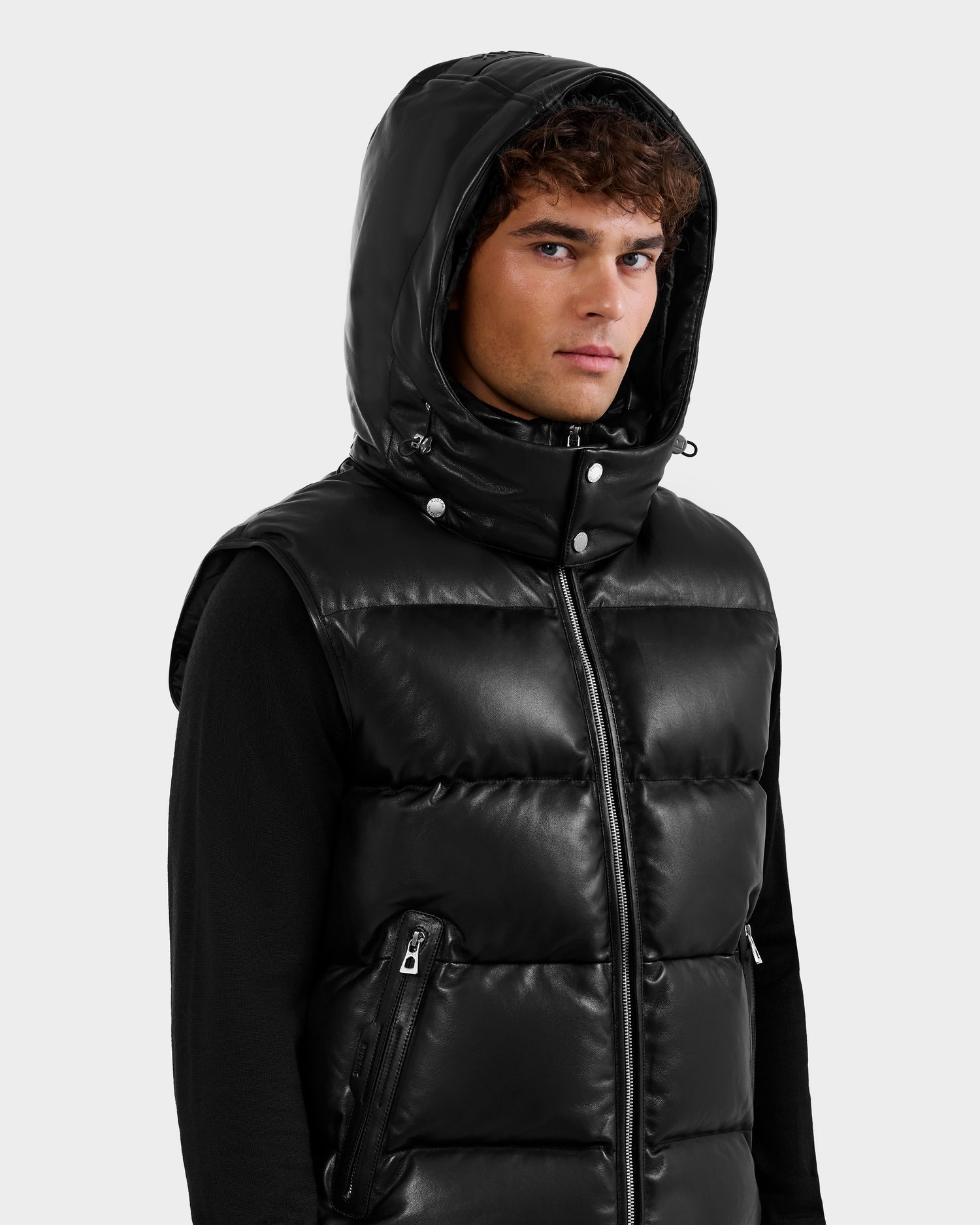A young man wears the Rudsak HENRY X - 6125679 Black shiny puffer vest with a hood and zippered pockets over a black long-sleeve shirt, gazing to the side against a plain light background.
