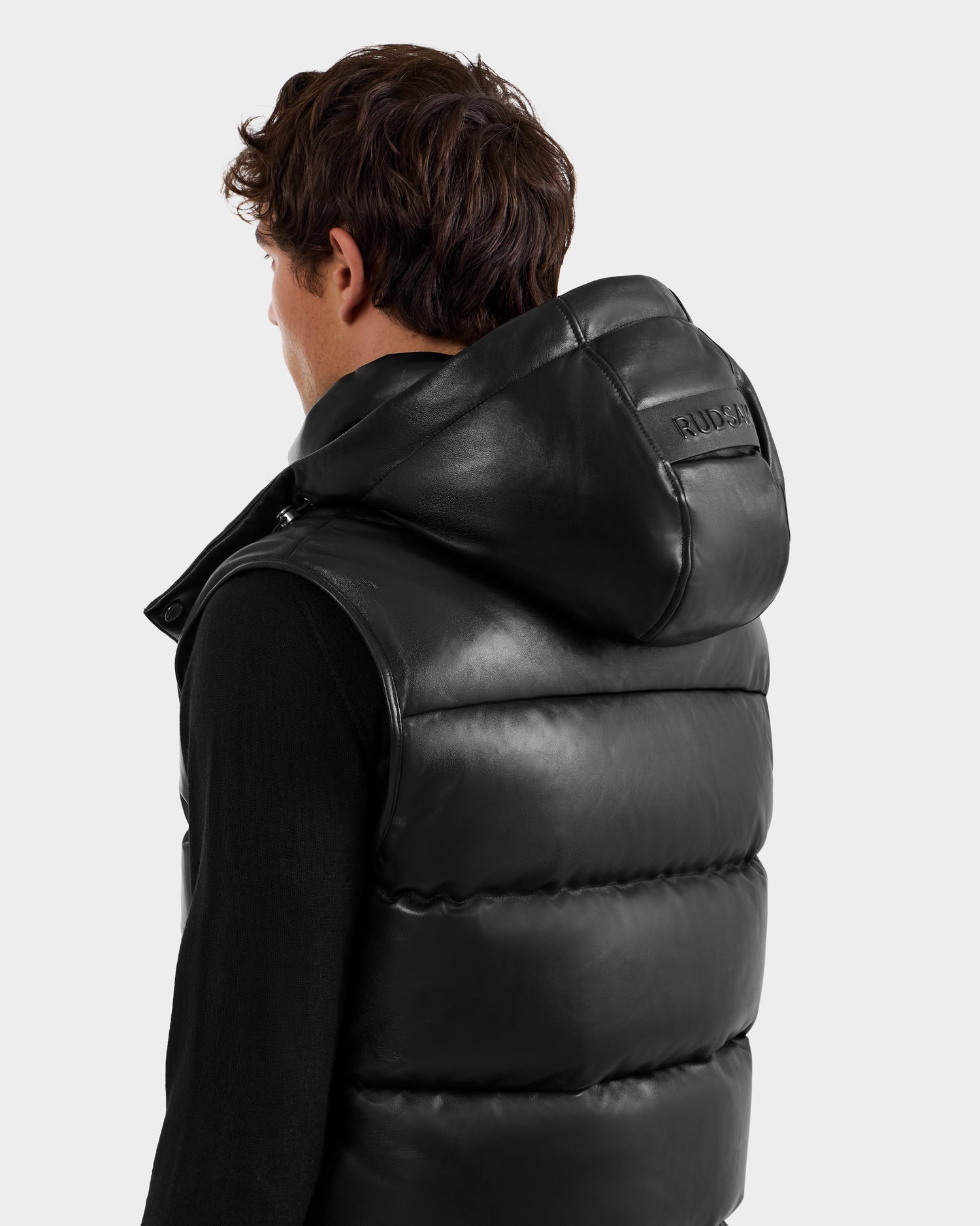 A person faces away from the camera wearing the Rudsak HENRY X - 6125679 Black padded leather vest with a large hood. The background is plain and light gray.