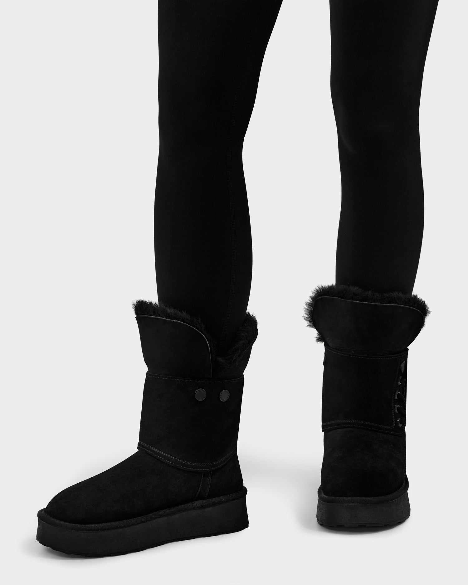 A person wears Rudsak's INESA - L225026 Black women’s suede platform boots with wool lining, fur trim, and button details, paired with black leggings, standing on a light gray background.