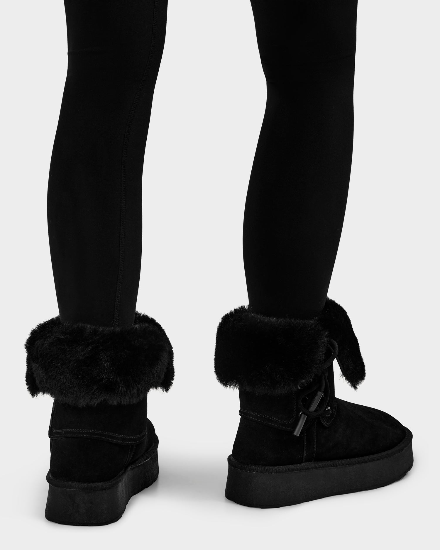 A person wearing black leggings and Rudsak INESA - L225026 Black women’s suede platform boots with wool lining and a sturdy sole is shown from the calves down, standing on a white background.