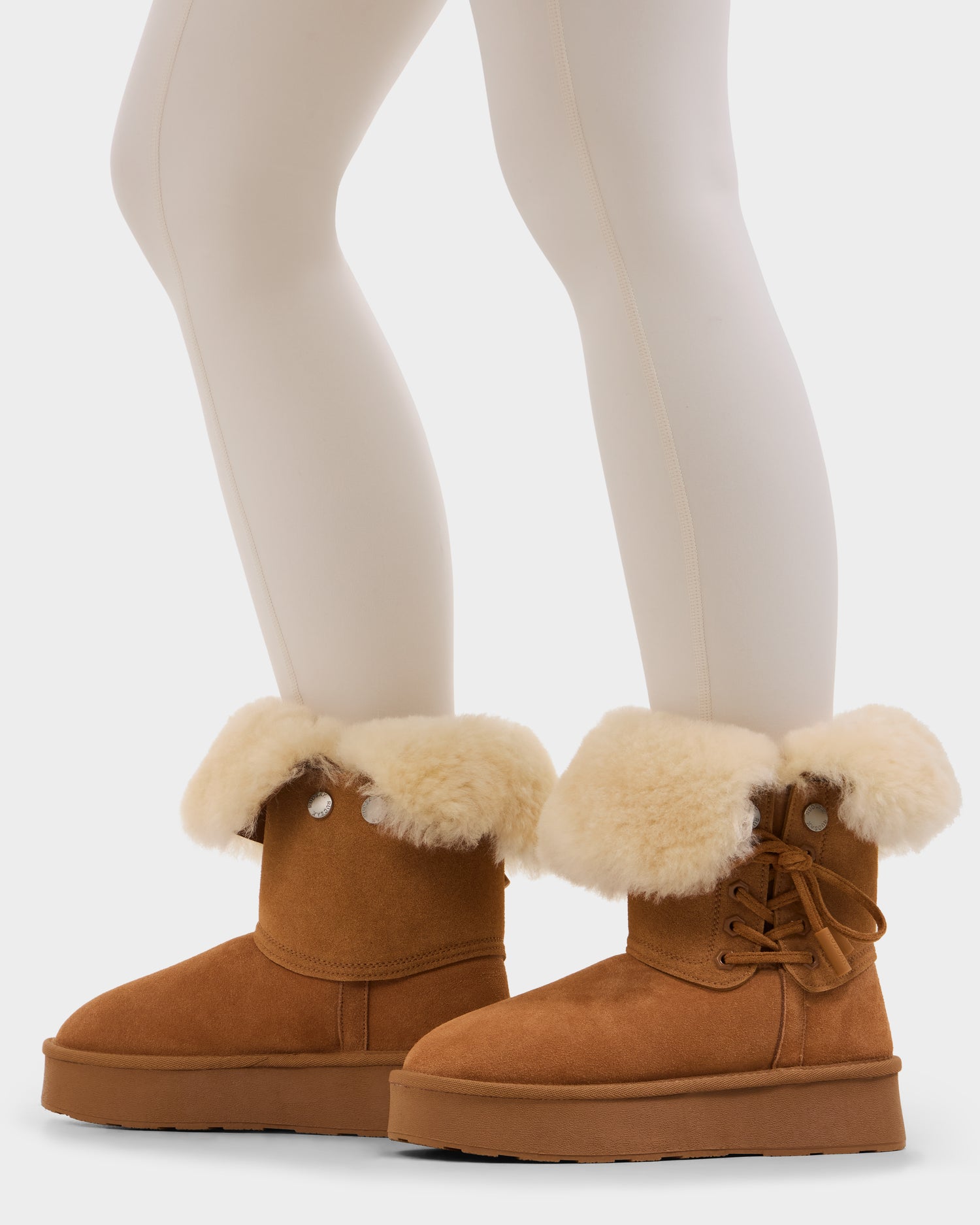 A person wears cream leggings and Rudsak’s INESA - L225026 Tan women’s suede platform boots featuring thick, folded-over shearling cuffs, wool lining, and side lace accents, standing on a white background.