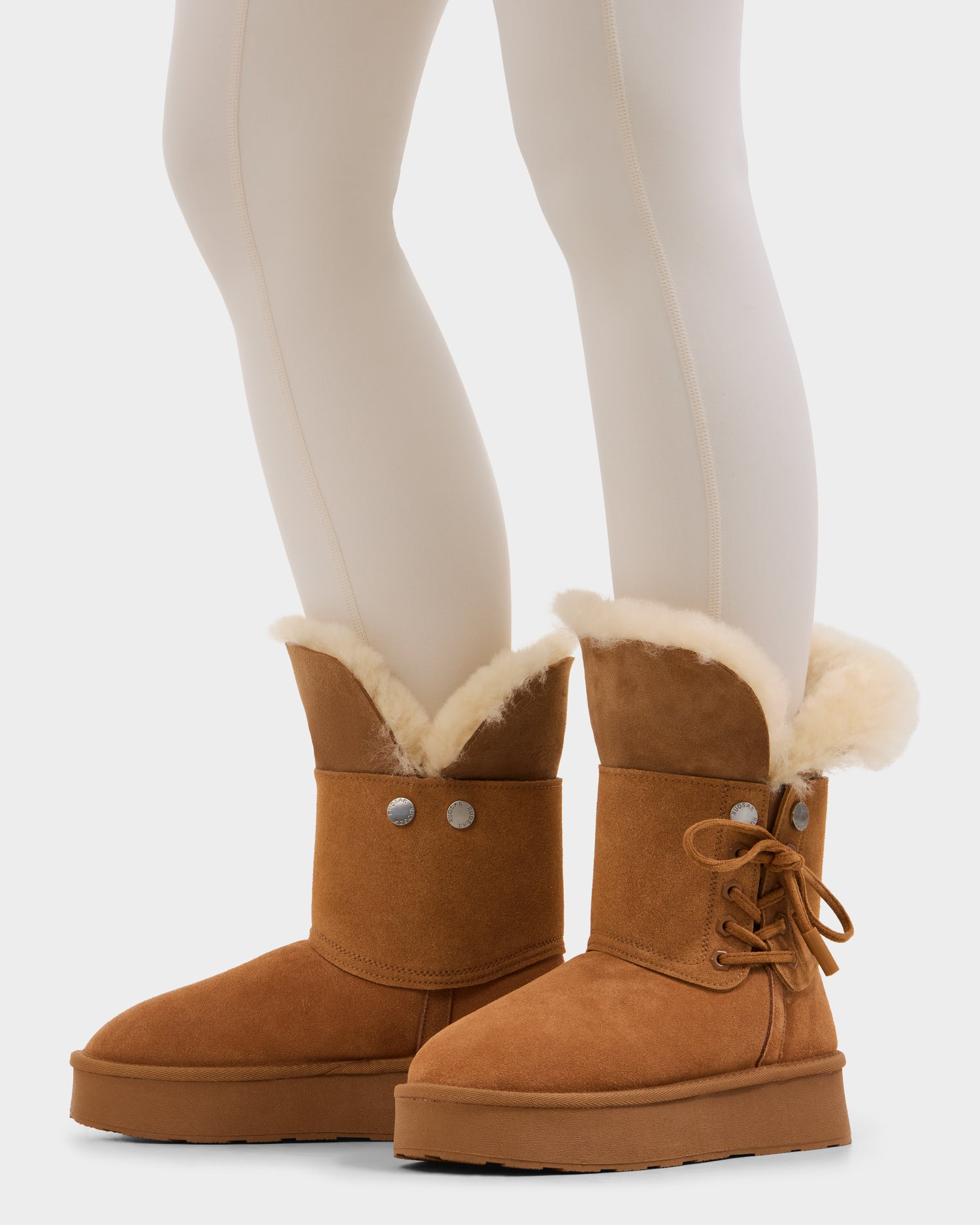 A person wearing cream-colored tights and Rudsak INESA - L225026 Tan women’s suede platform boots with wool lining, button, and lace details stands against a plain white background.
