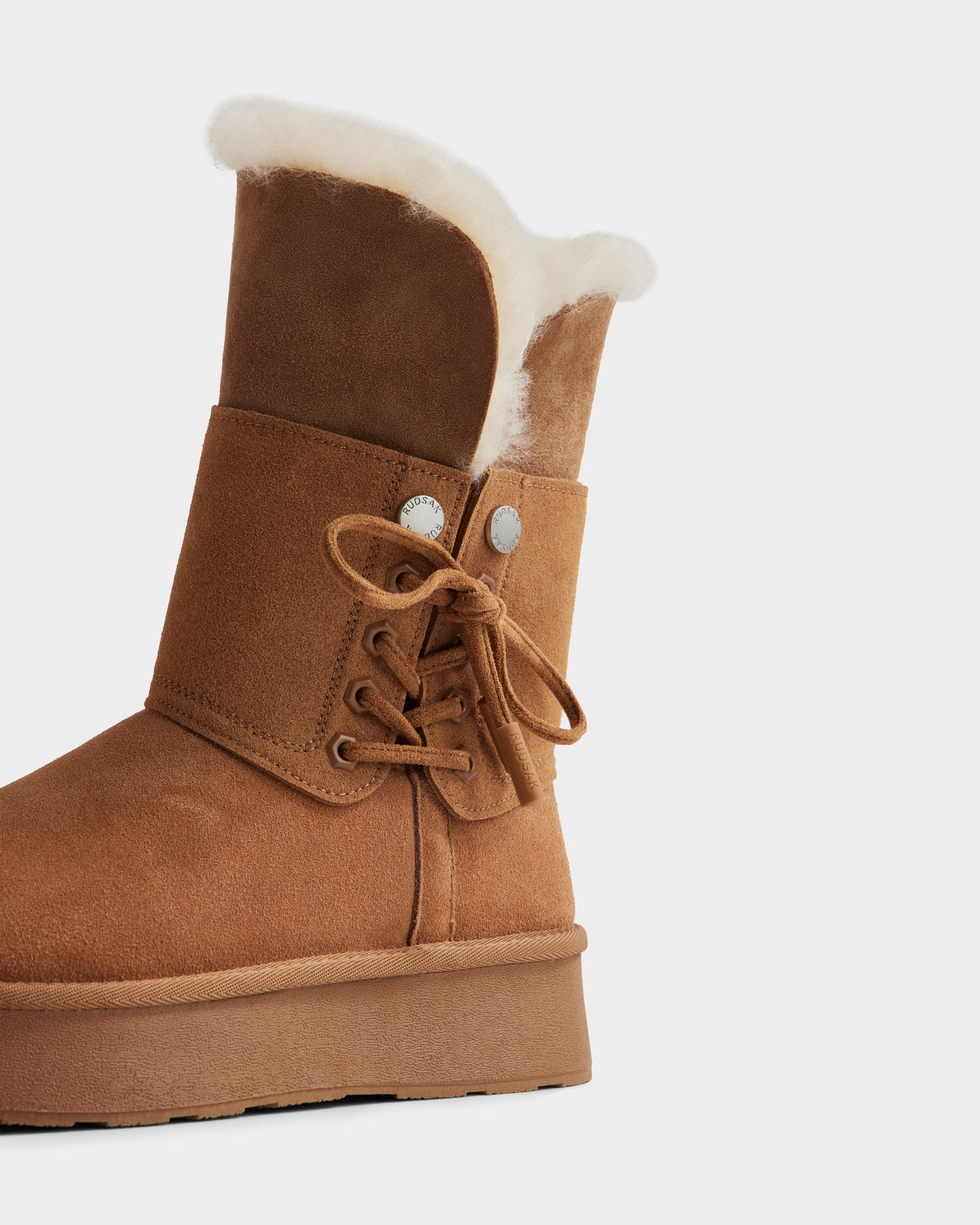 The INESA - L225026 Tan by Rudsak is a brown women’s suede platform boot with white wool lining, side bow laces, metal eyelets, and a lightweight sole. Only the right boot is partially visible against a white background.