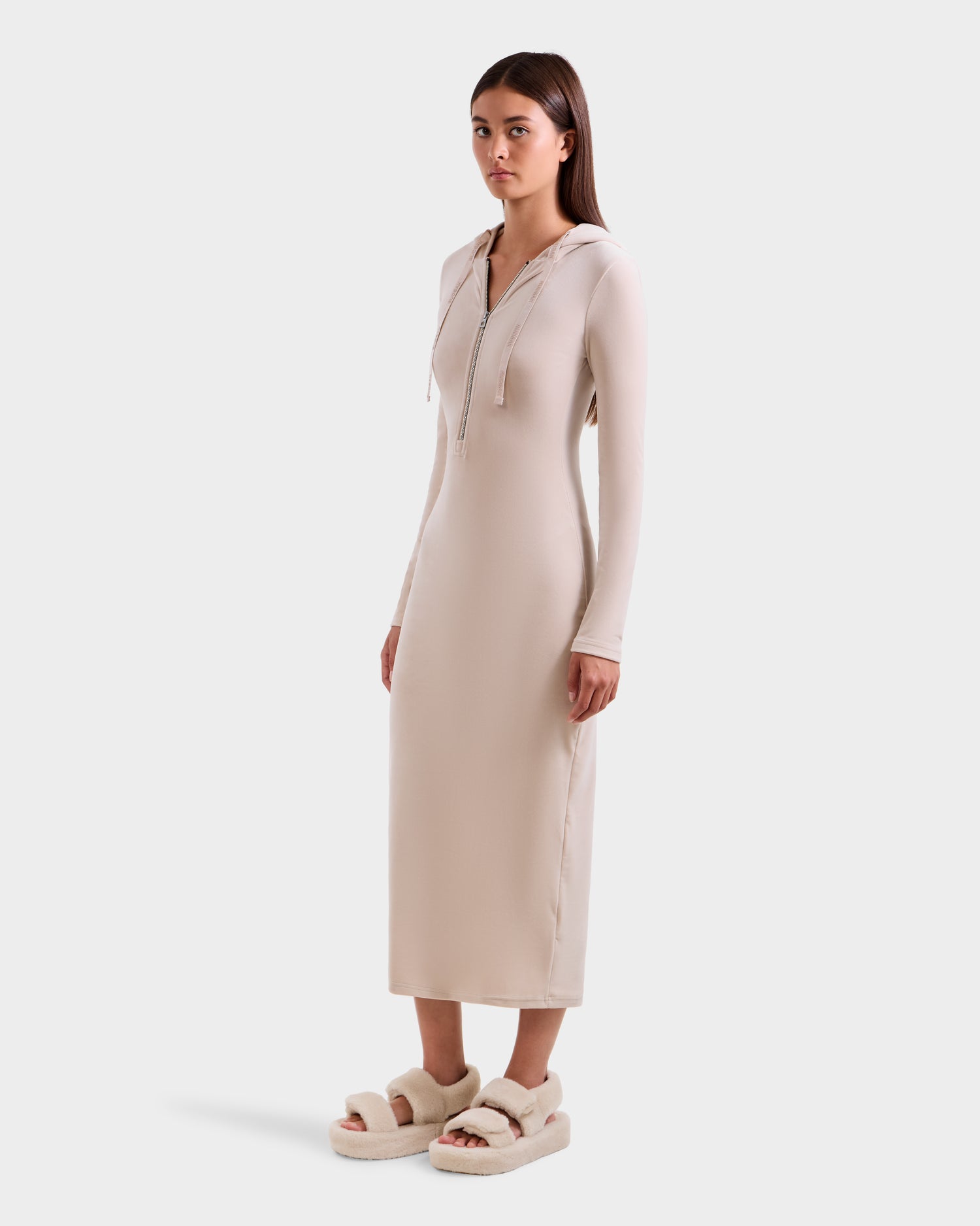 A woman stands in the Rudsak ISABELLA - L525502 Cream long fitted velour hooded dress with a front zipper and long sleeves, paired with beige platform fuzzy slippers, facing slightly to the side against a plain light background.