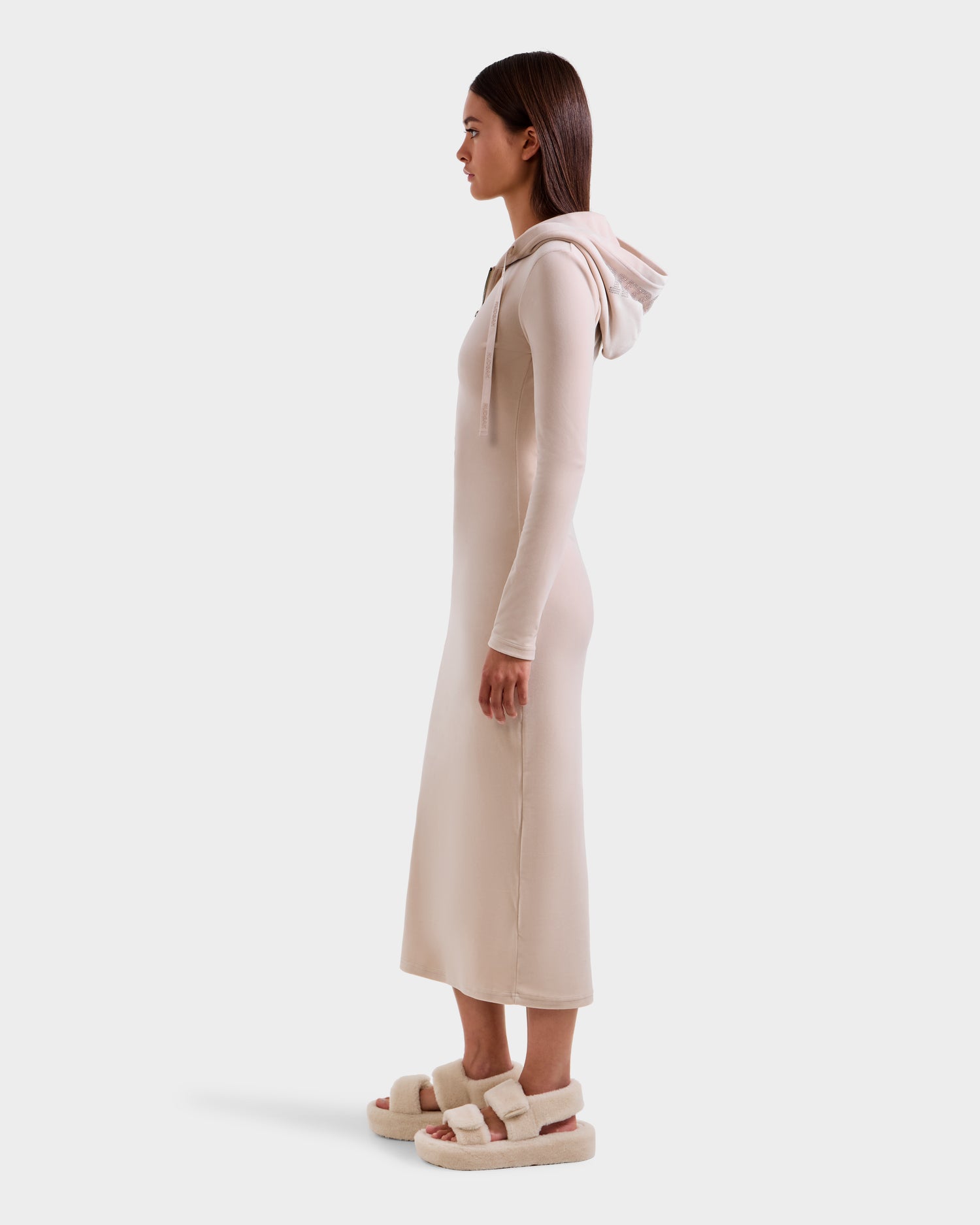 A woman stands in profile wearing the Rudsak ISABELLA - L525502 Cream velour hooded dress with long sleeves and beige fuzzy sandals against a plain white background.
