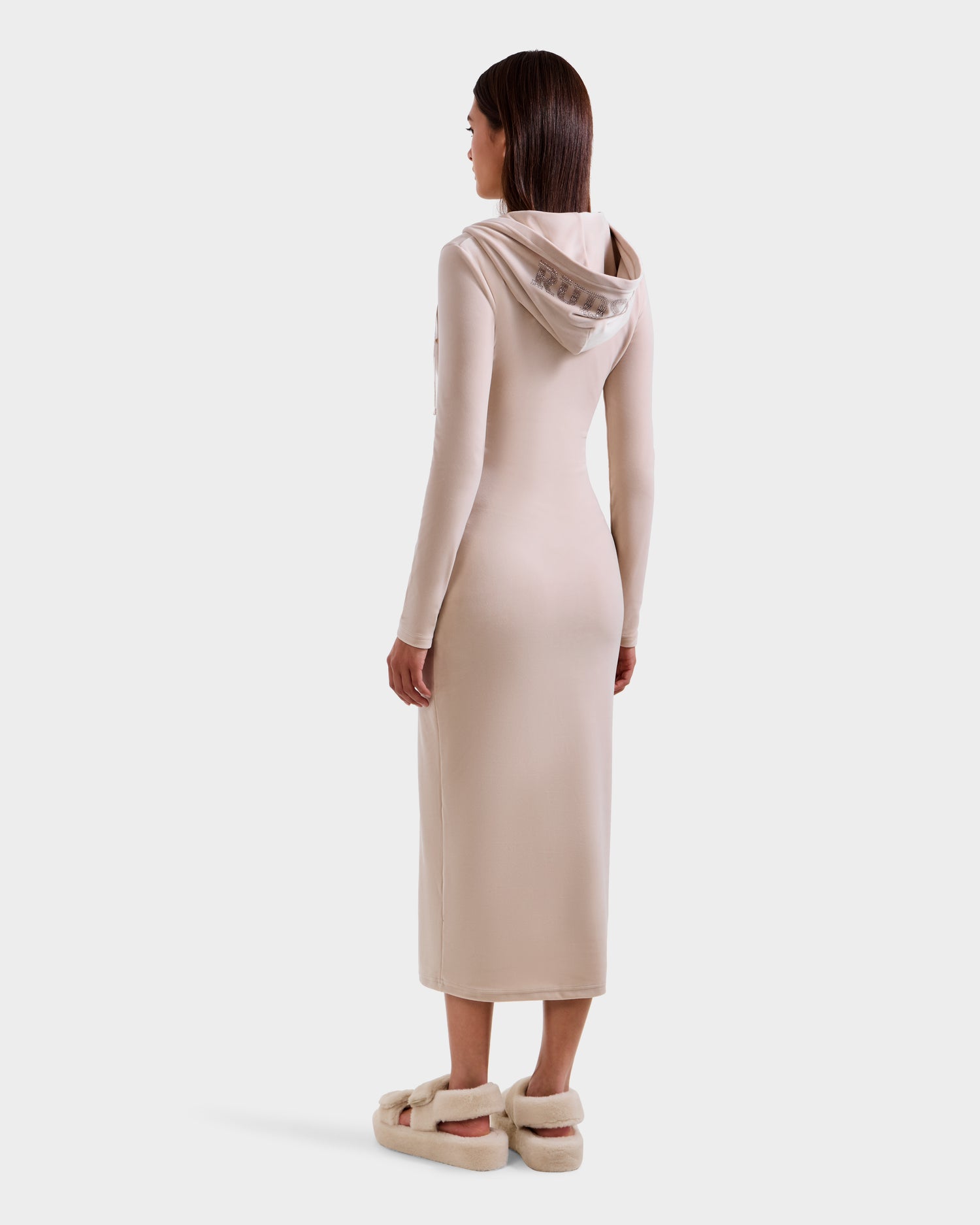 A woman with straight brown hair stands facing away, wearing the Rudsak ISABELLA - L525502 Cream velour hooded dress with long sleeves and beige fuzzy slippers against a plain white background.