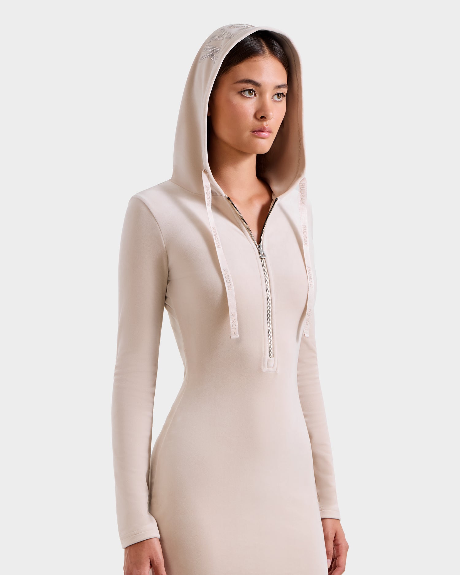 A woman wears the ISABELLA - L525502 Cream fitted long-sleeve velour hooded dress by Rudsak, featuring a front zipper. She stands facing slightly right against a plain light background, hood up and expression neutral.