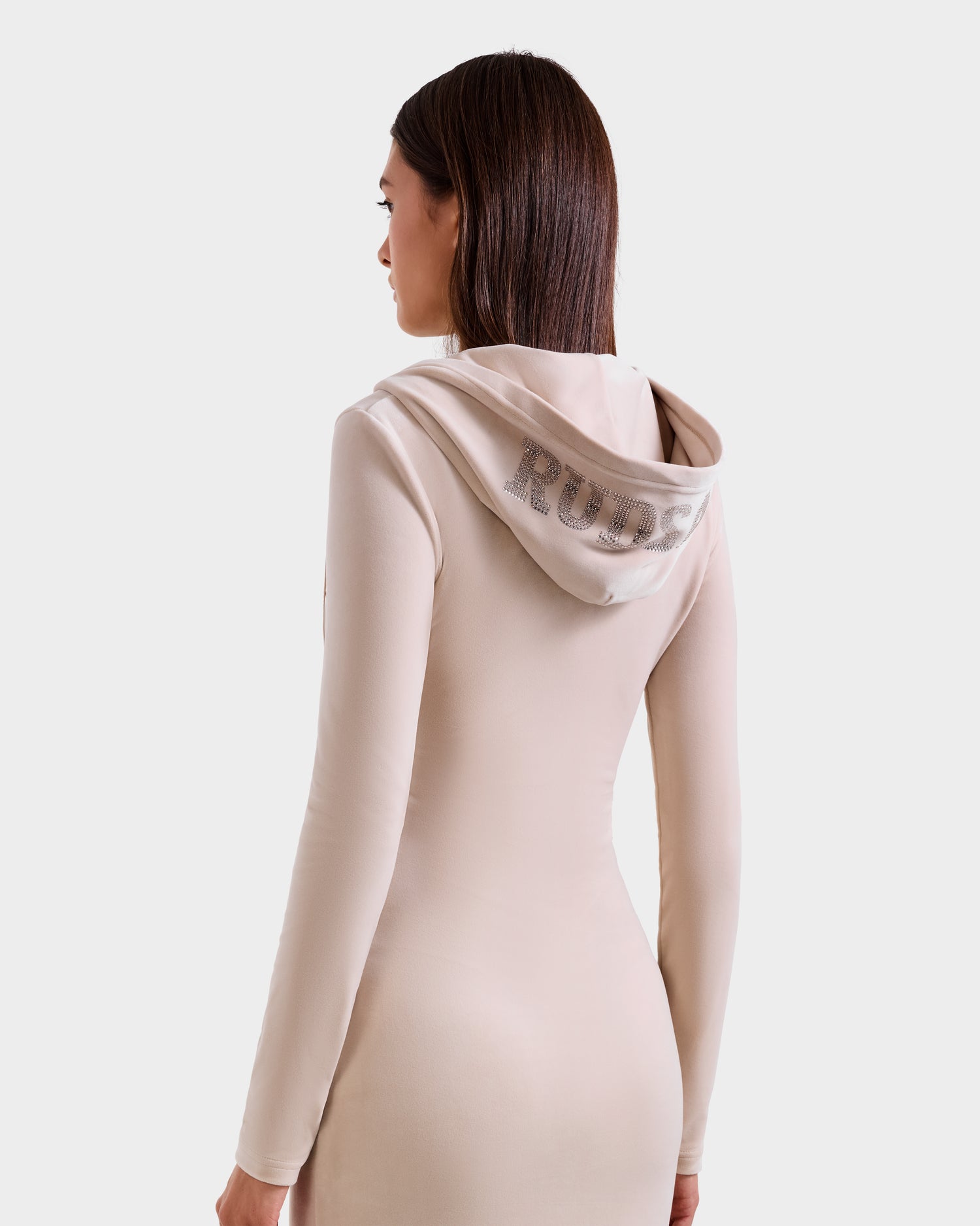 A woman with straight brown hair models the Rudsak ISABELLA - L525502 Cream velour hooded dress. She is turned slightly away, highlighting the rhinestone “Rude” on the back. The background is plain light gray.