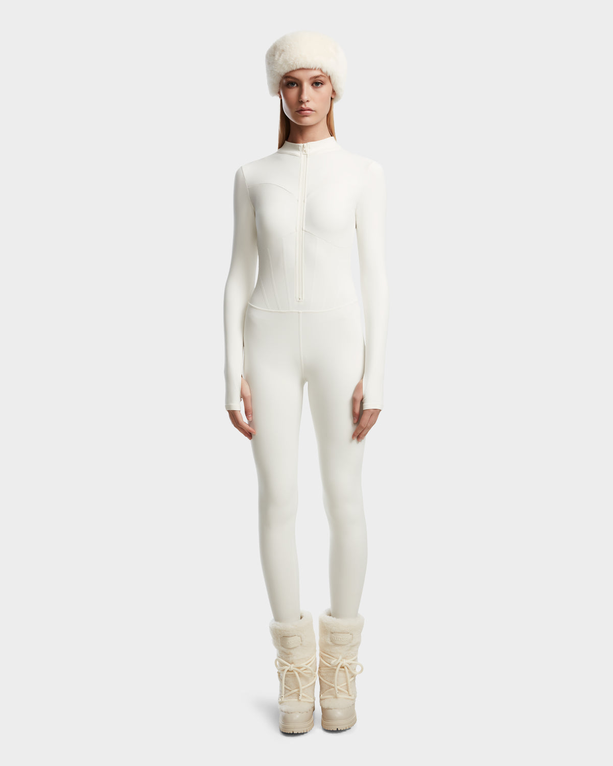A woman wears the Rudsak IVY - 8524599 Iceland fitted all-white zip-up ski suit, paired with a white fur hat and beige snow boots, exuding timeless ski-chic style against a plain light background.