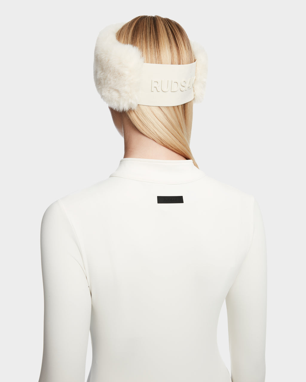 A person with straight blonde hair models the IVY - 8524599 Iceland women’s stretch bodysuit by Rudsak and white earmuffs, shown from the back against a plain light background for a sleek, ski-chic look.