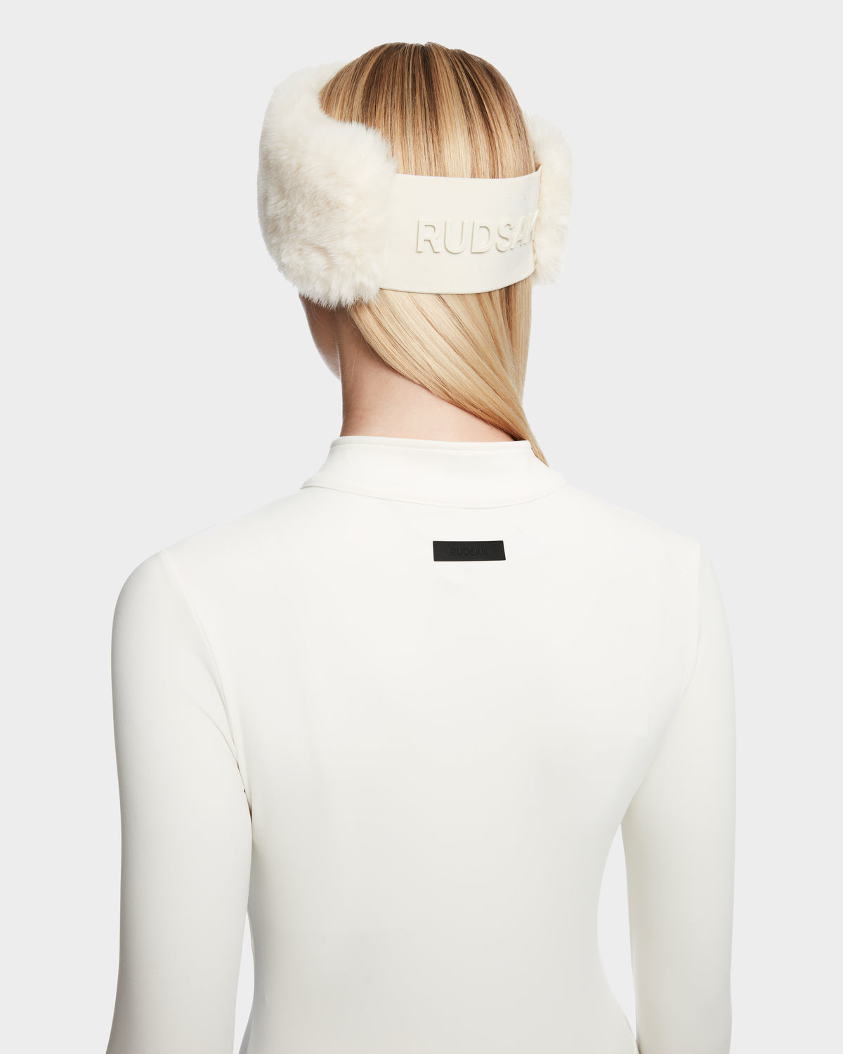 A person with straight blonde hair models the IVY - 8524599 Iceland women’s stretch bodysuit by Rudsak and white earmuffs, shown from the back against a plain light background for a sleek, ski-chic look.