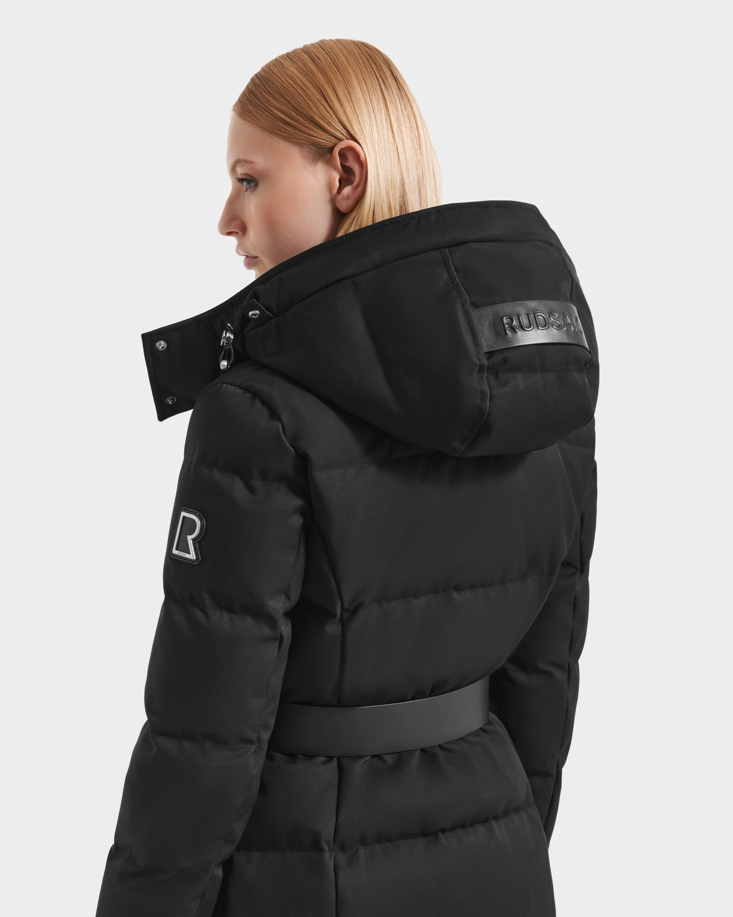 A woman with straight blonde hair models the Rudsak IZABELLA H - 8125644 Black puffer jacket, featuring a belt, a hood with the Rudsak logo, and an R patch on the sleeve. She is shown from the back and side.