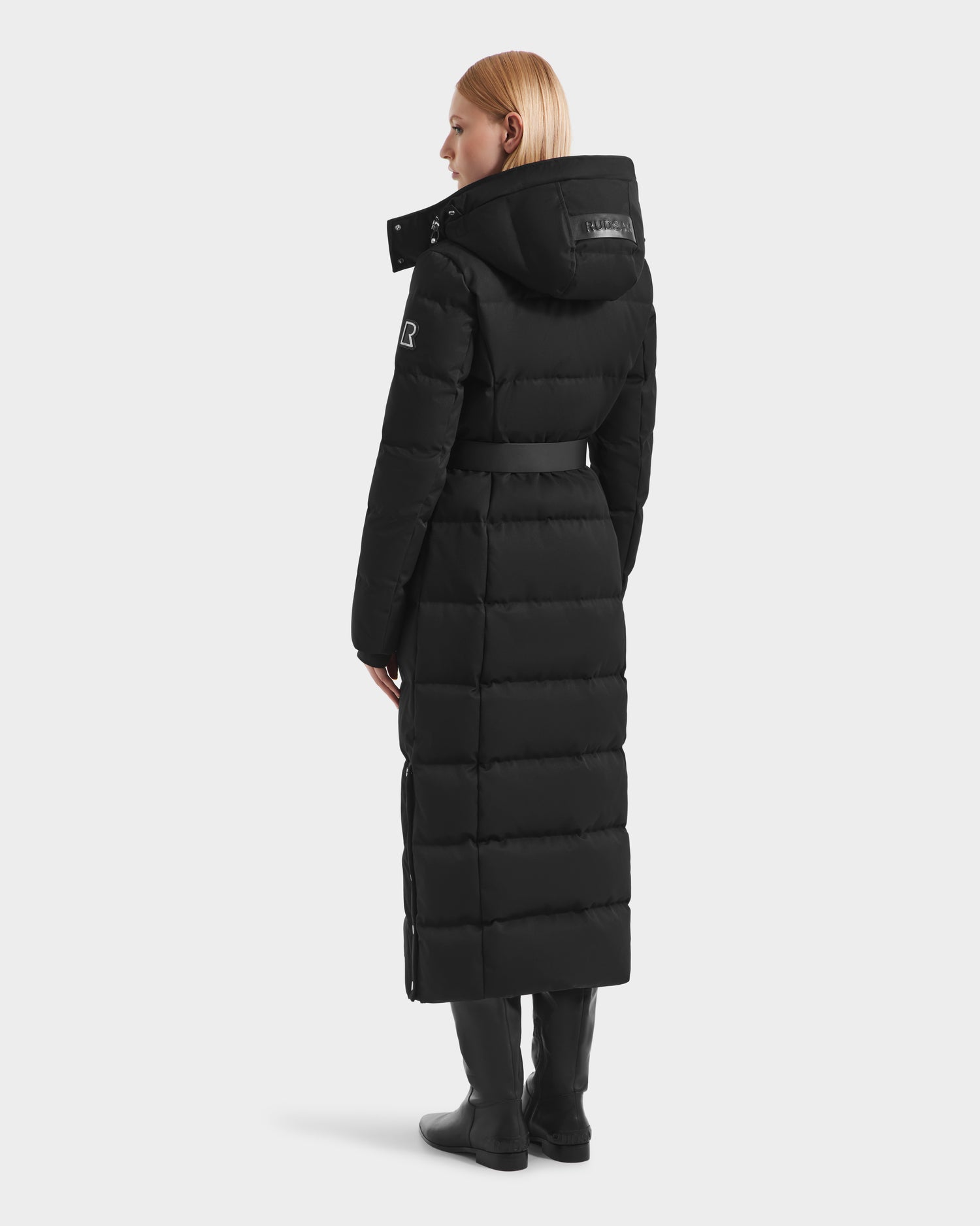 A person with straight blonde hair wears the Rudsak IZABELLA H - 8125644 Black women’s long puffer coat with a hood and belt, paired with black boots. They stand facing away on a plain white background.
