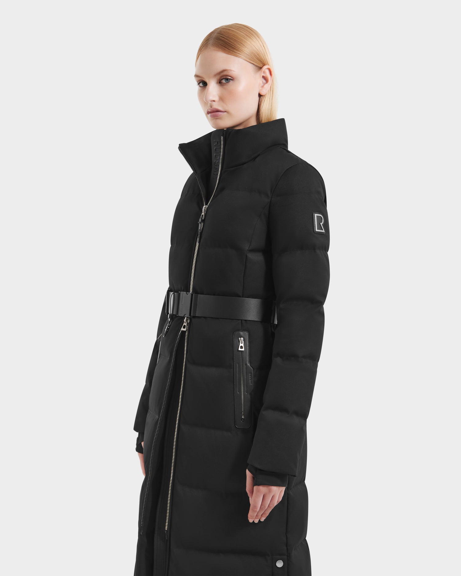A woman with straight blonde hair models the Rudsak IZABELLA H - 8125644 Black long women's puffer winter jacket with a high collar and zippered pockets, standing against a light gray background.