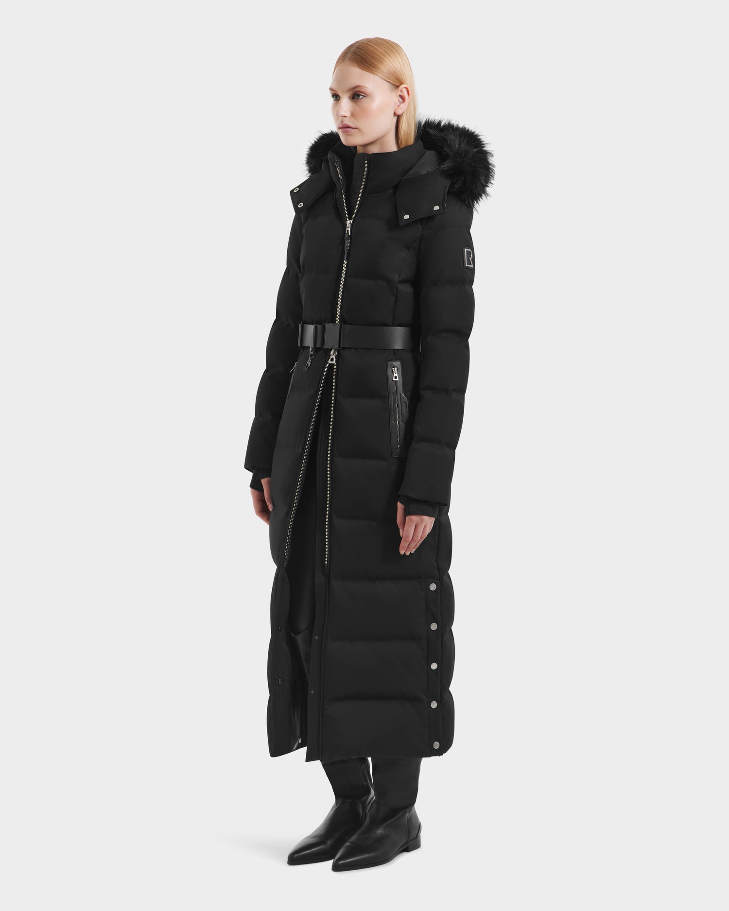 A woman wears the Rudsak IZABELLA H - 8125644 Black puffer coat, featuring a fur-trimmed hood, belted waist, and snap buttons on the sides. She pairs this stylish winter jacket with black pants and ankle boots.
