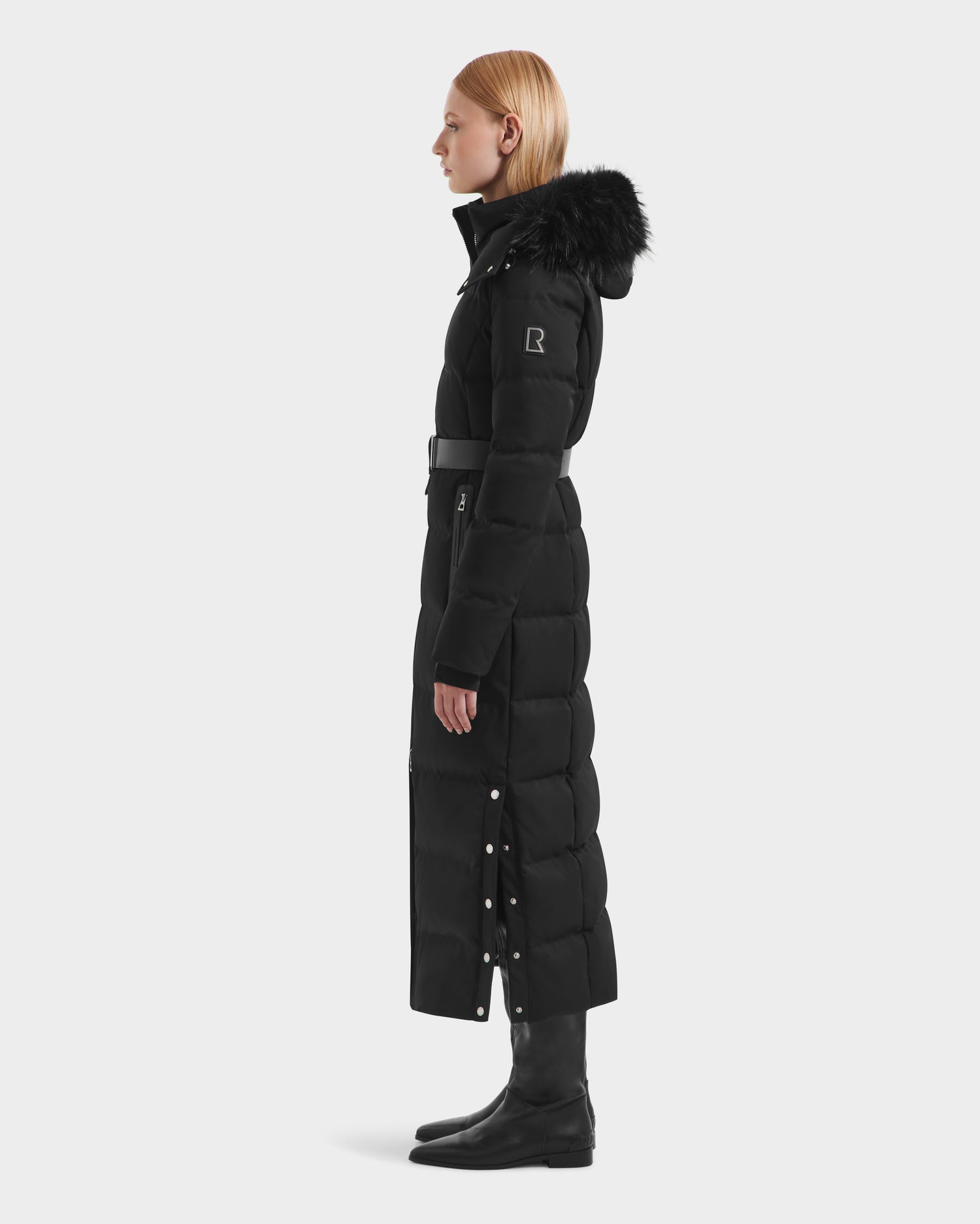 A woman with straight blonde hair models the Rudsak IZABELLA H - 8125644 Black puffer coat, featuring a belted waist and fur-trimmed hood, paired with tall black boots, standing in profile against a white background.