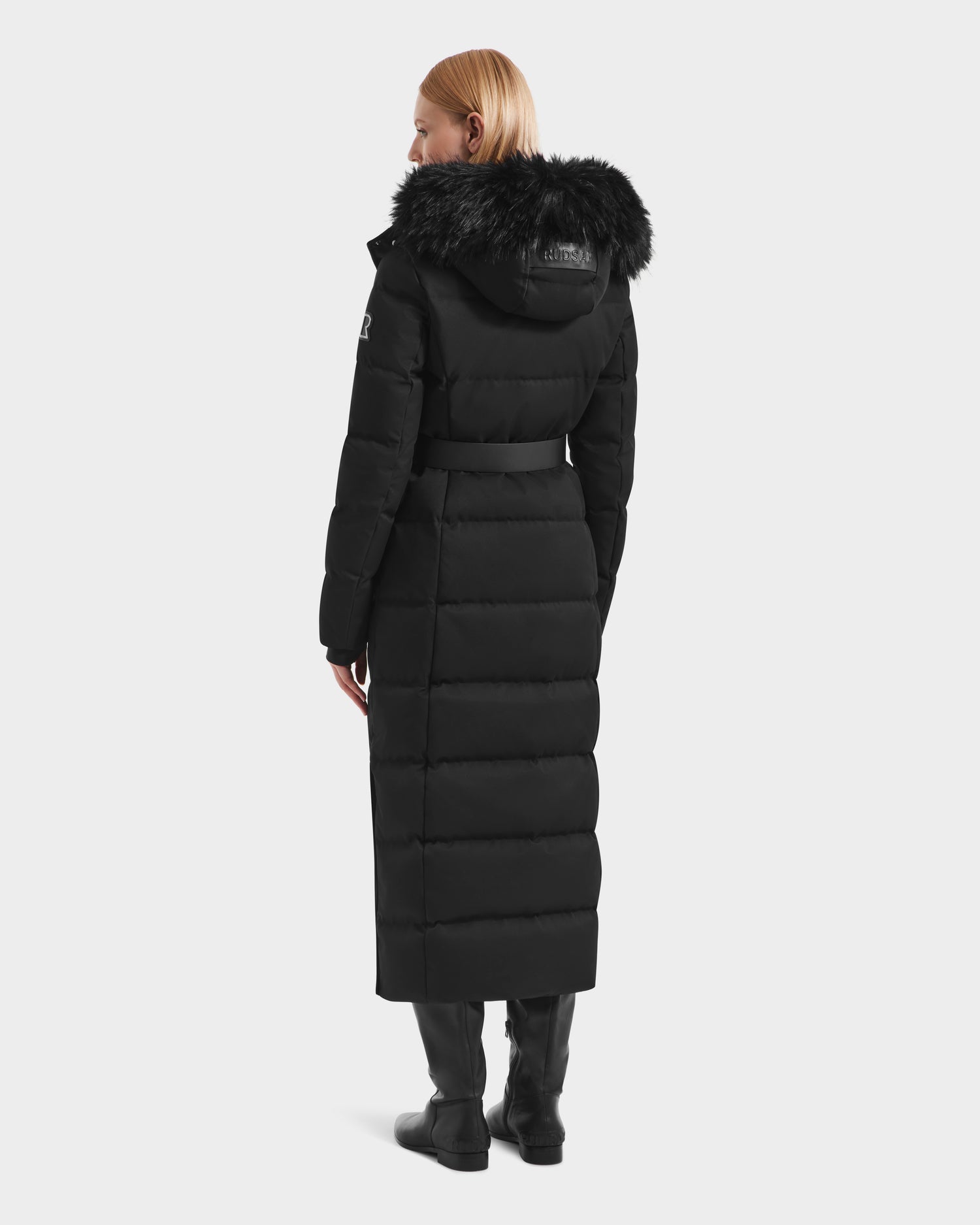 A person with light hair is wearing the Rudsak IZABELLA H - 8125644 Black women's puffer winter jacket with a fur-lined hood, along with black pants and black boots, standing facing away from the camera against a plain background.