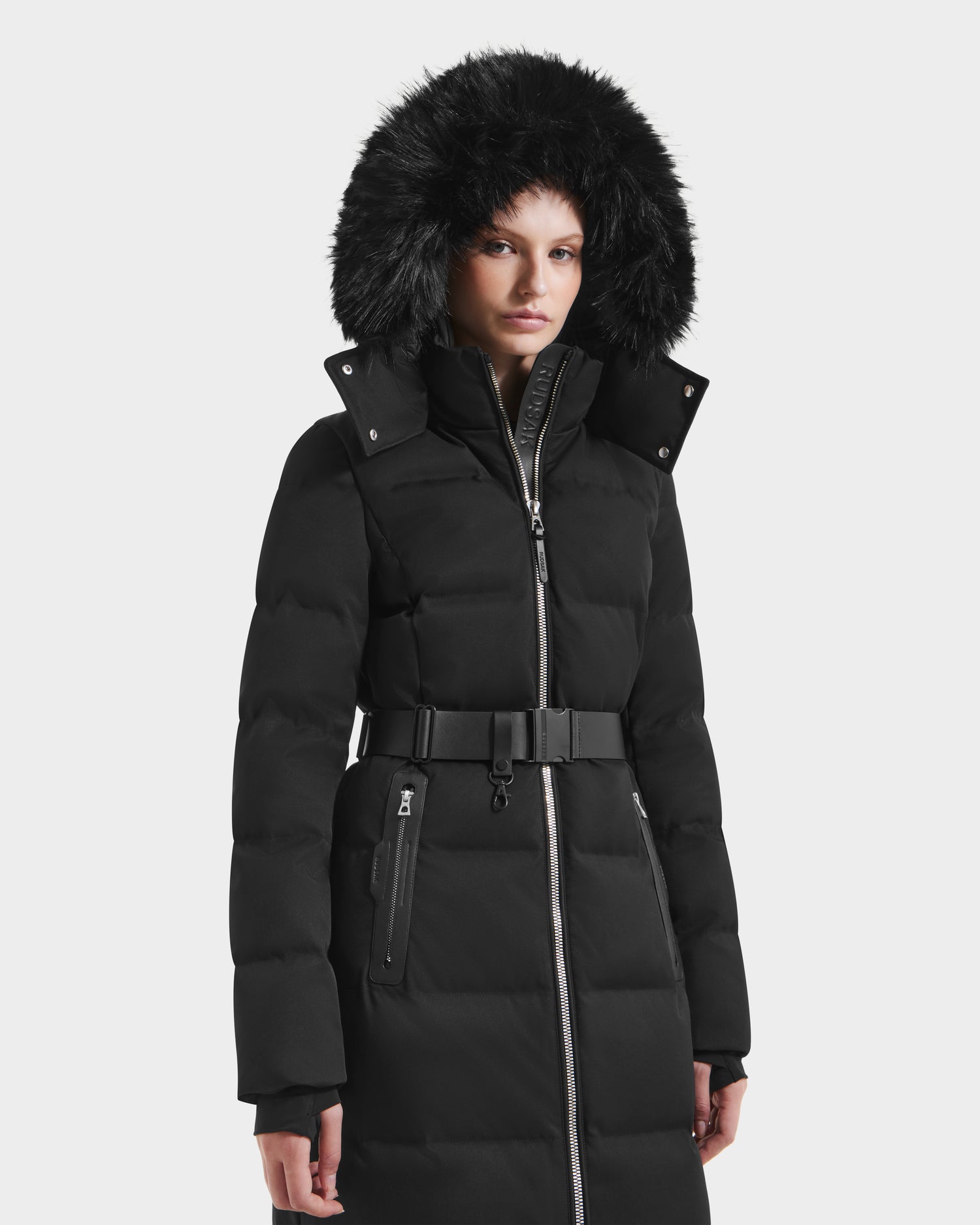A woman models the Rudsak IZABELLA H - 8125644 Black long puffer with a belted waist and furry hood against a white background—a chic winter jacket.