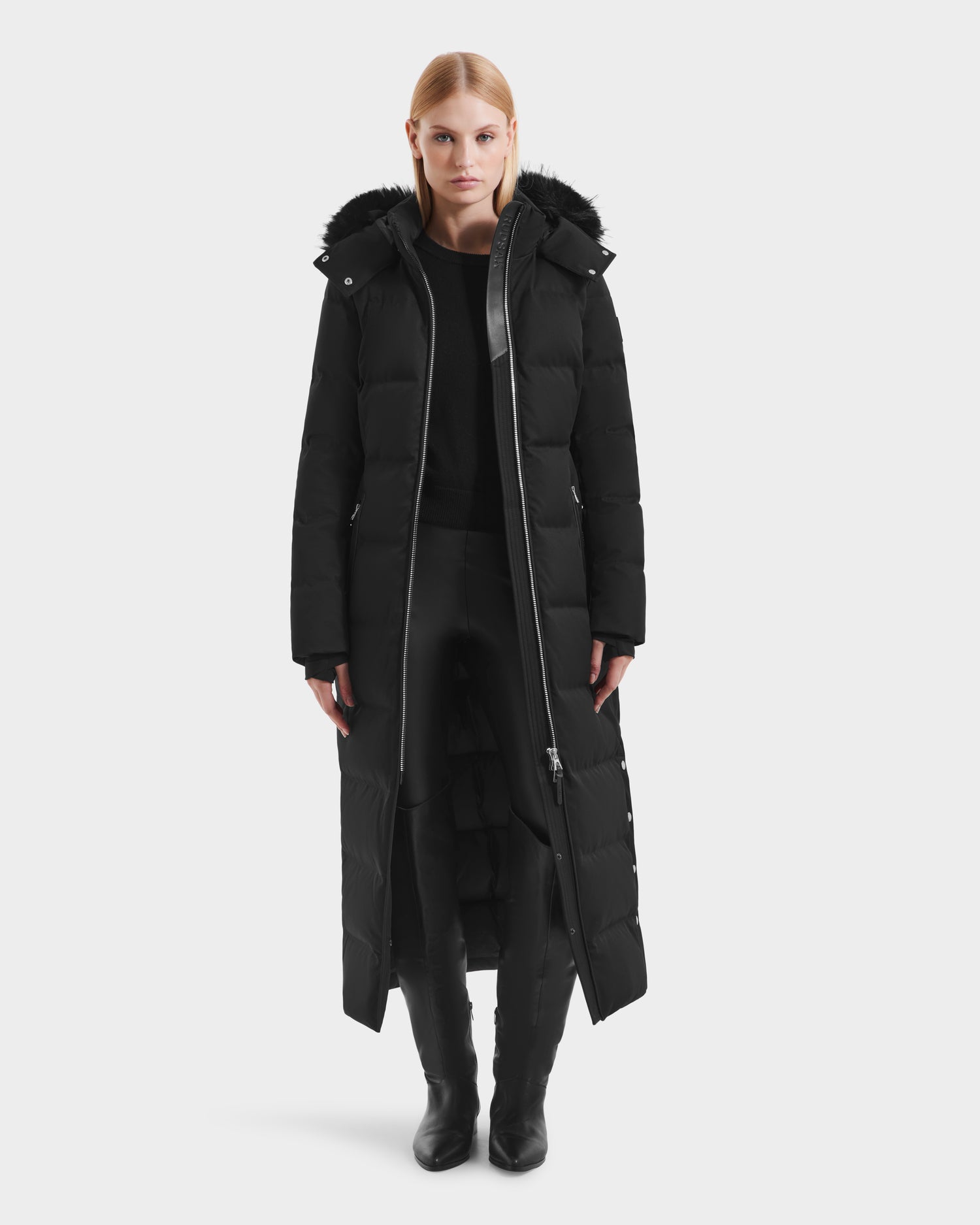 A woman stands facing forward against a white background, wearing the Rudsak IZABELLA H - 8125644 Black puffer coat with a hood, paired with black pants and black boots.