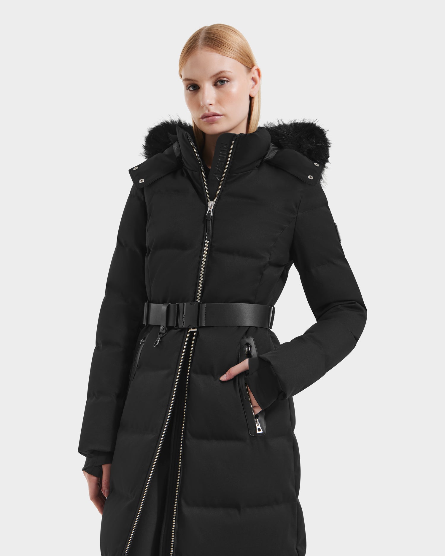 A person wears the Rudsak IZABELLA H - 8125644 Black puffer coat, featuring a belted waist, zippered front, and black faux fur-trimmed hood, standing against a plain light background.