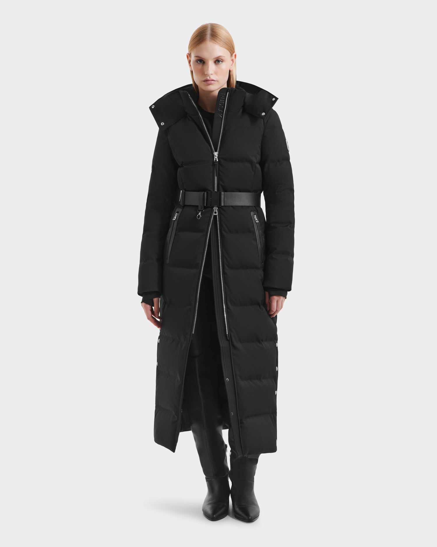 A woman with straight blonde hair wears the Rudsak IZABELLA H - 8125644 Black puffer coat, featuring a belt, zippered front, and high collar, paired with black boots, standing against a plain light background.
