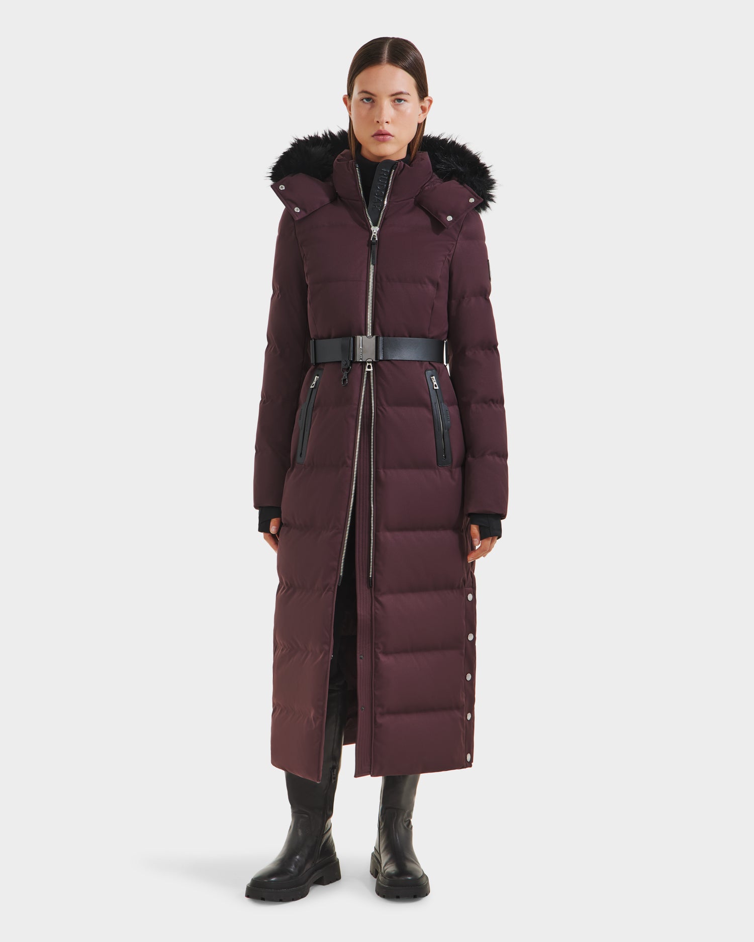 A woman faces forward wearing the Rudsak IZABELLA H - 8125644 Bordeaux long plum puffer with down fill, fur-trimmed hood, black belt, zipper and snap closures, black boots, and gloves against a plain white background.